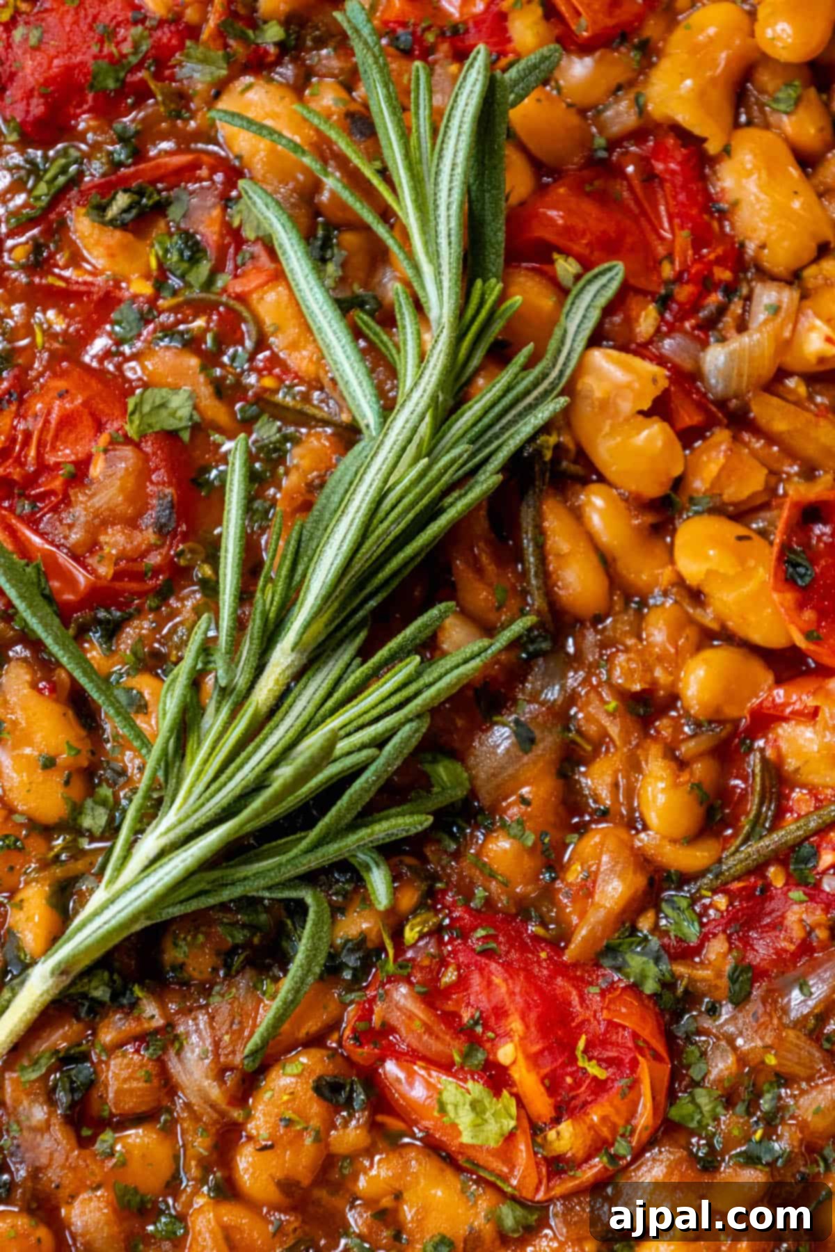 A vibrant brothy dish featuring tender white beans and ripe tomatoes, perfectly infused with a sprig of fresh rosemary, highlighting the fresh ingredients and rich flavors of the Mediterranean.