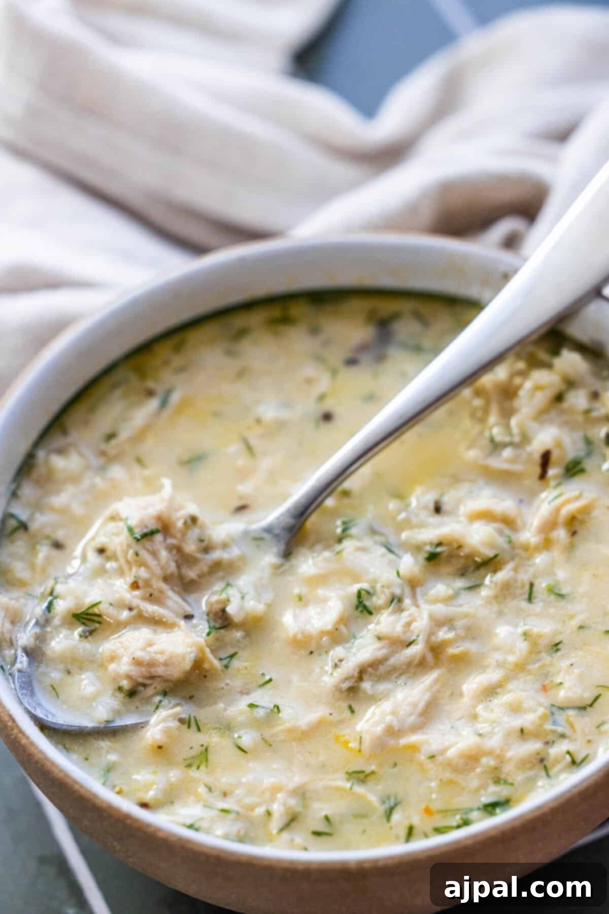 Velvety Greek Avgolemono 2 A bowl of creamy Greek avgolemono soup with herbs, shredded chicken, and a spoon resting inside. A beige cloth is visible in the background.
