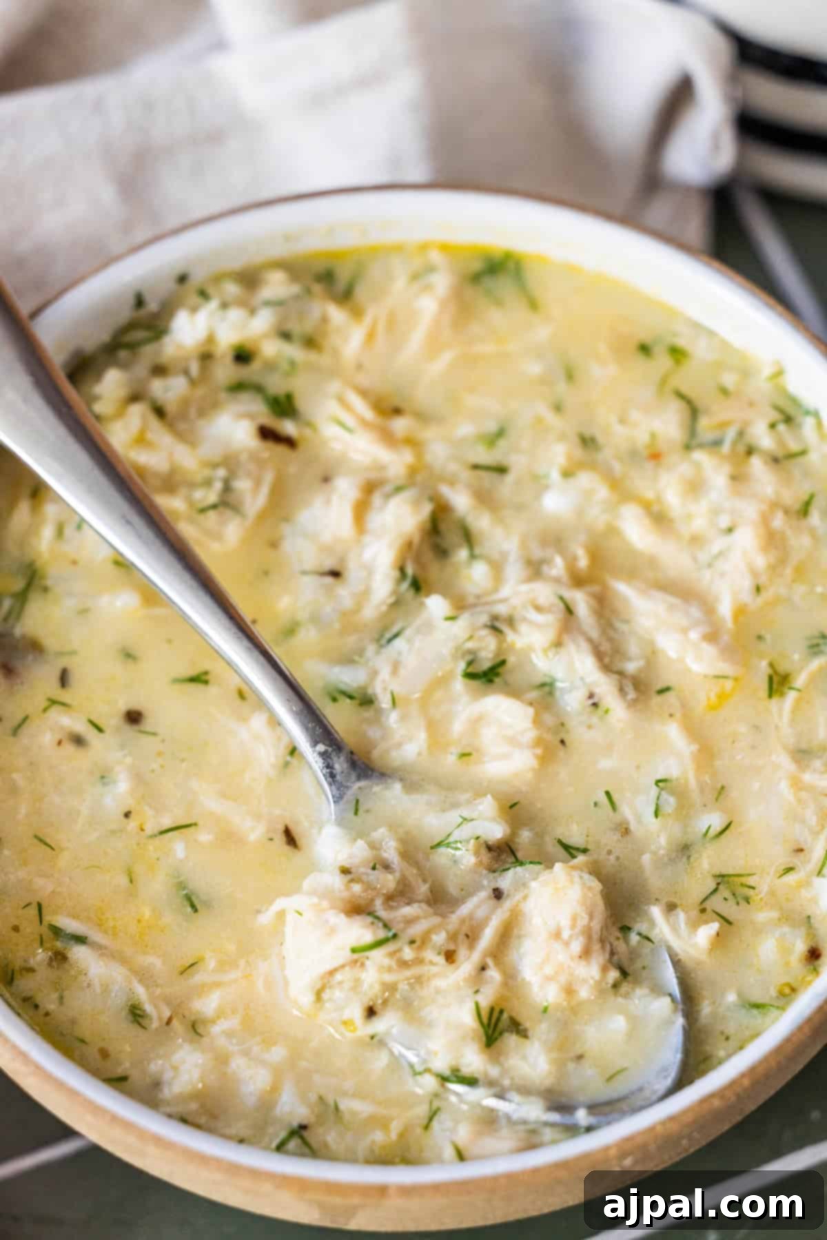 Velvety Greek Avgolemono 4 A bowl of creamy Greek avgolemono soup with herbs, featuring shredded chicken and a spoon resting inside the bowl.