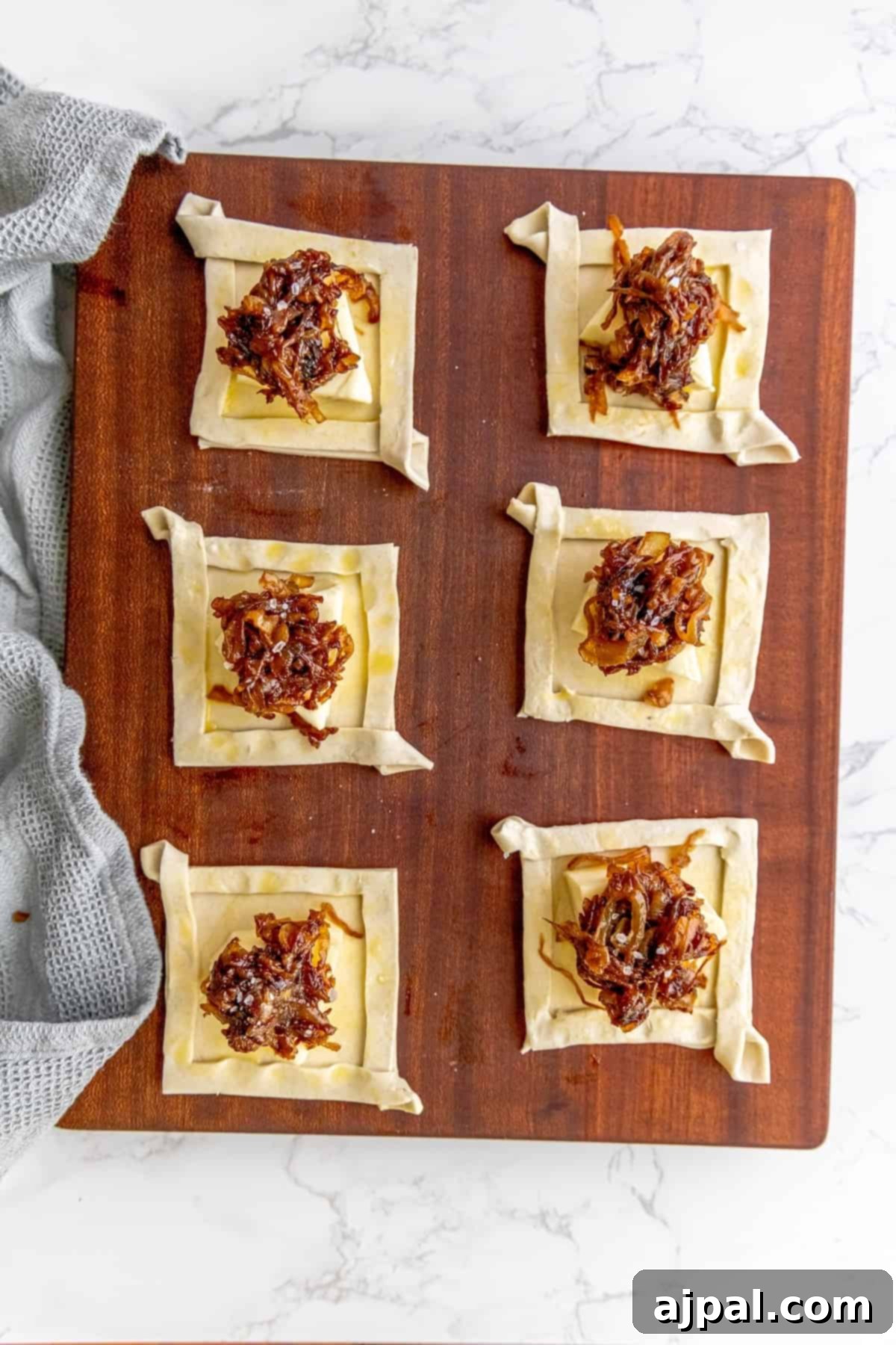 A wooden cutting board holds a tray of unbaked pastries, each generously topped with brie cheese and caramelized onions.