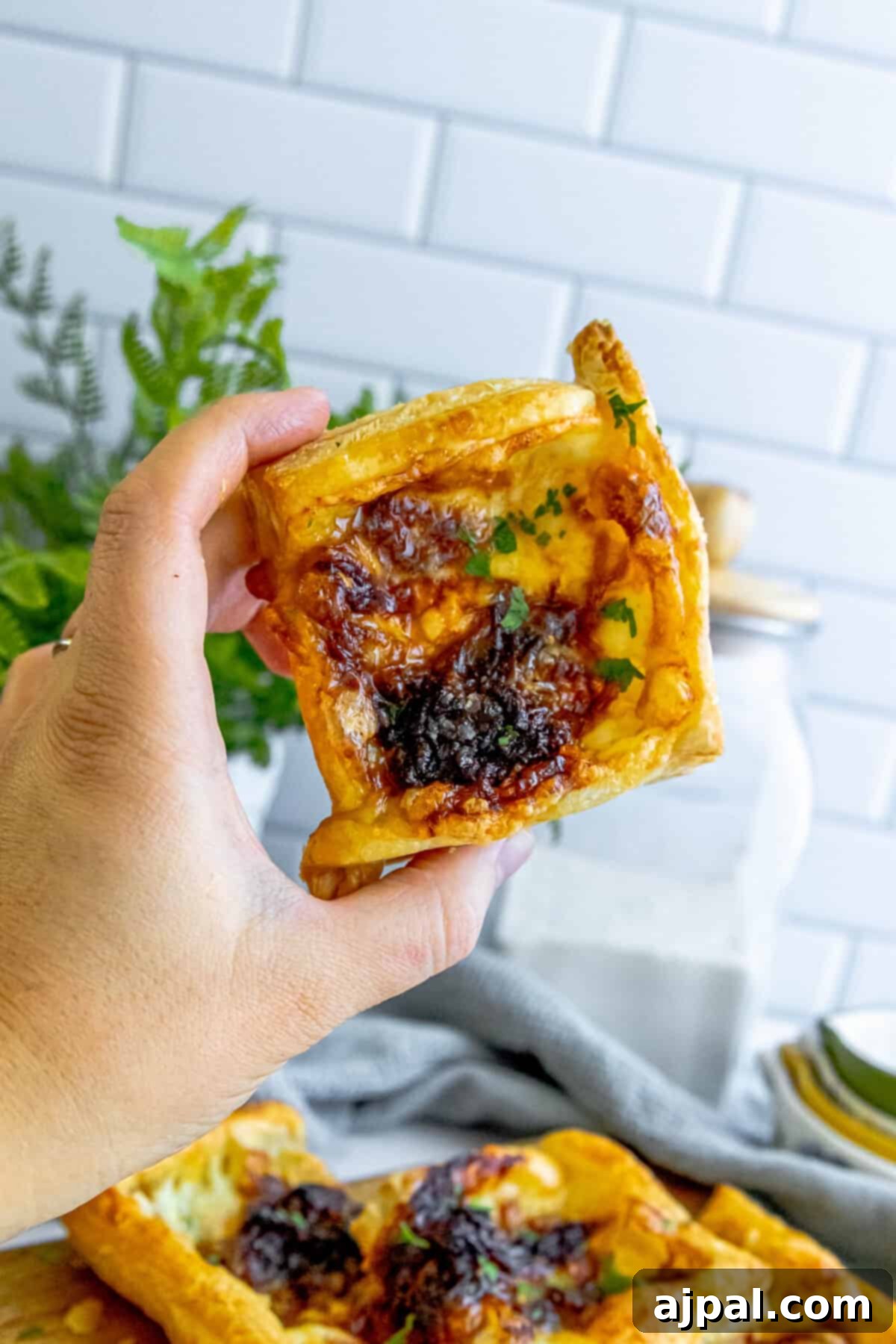 A hand gently holds a golden, flaky caramelized onion brie tart.