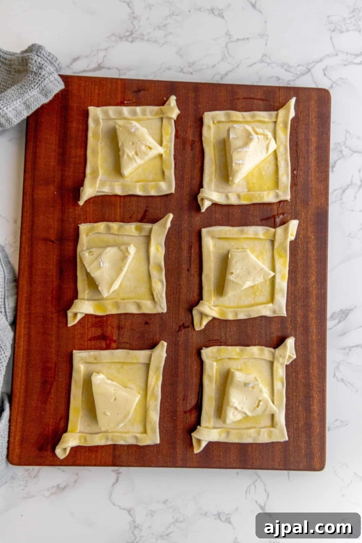 A wooden cutting board displaying several puff pastry squares, each with a piece of brie cheese placed in its center.