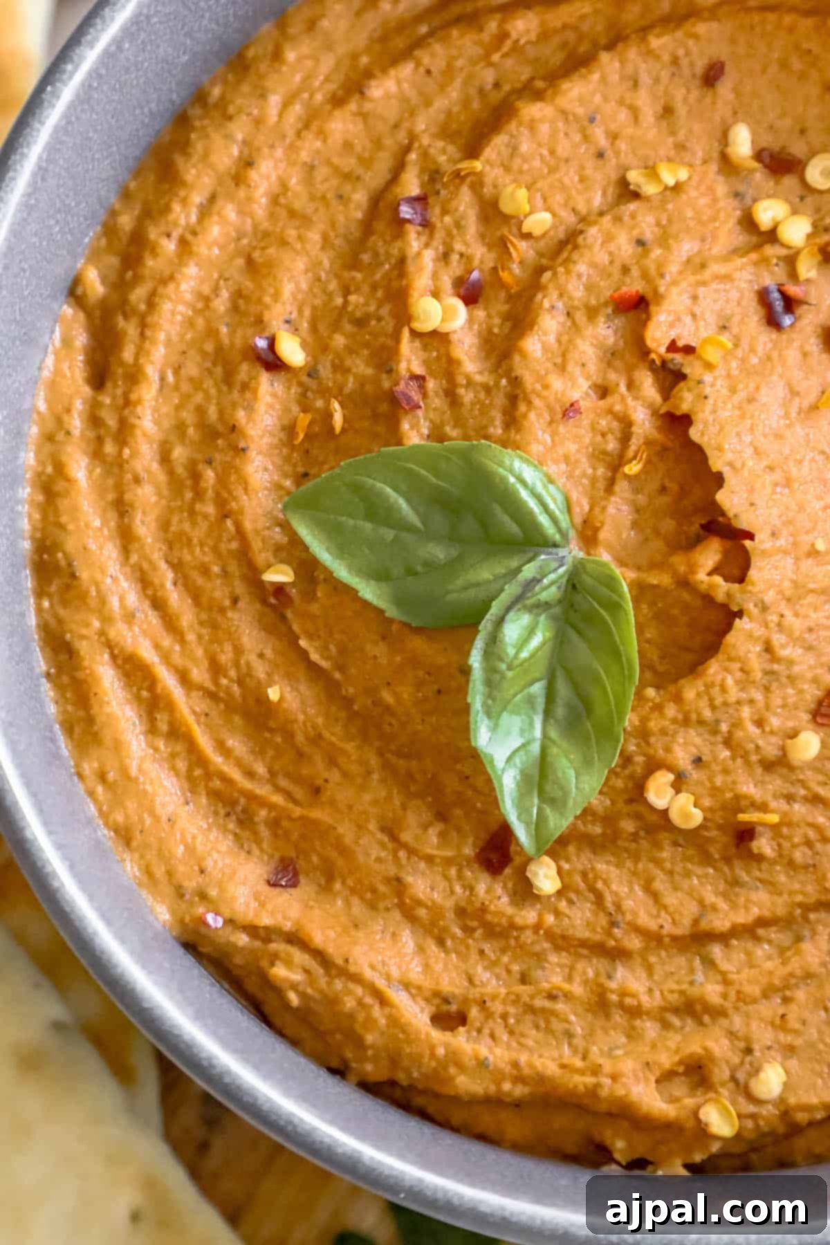 A beautifully presented bowl of smooth, red-hued pizza hummus, elegantly garnished with a fresh green basil leaf, resting on a rustic wooden cutting board.