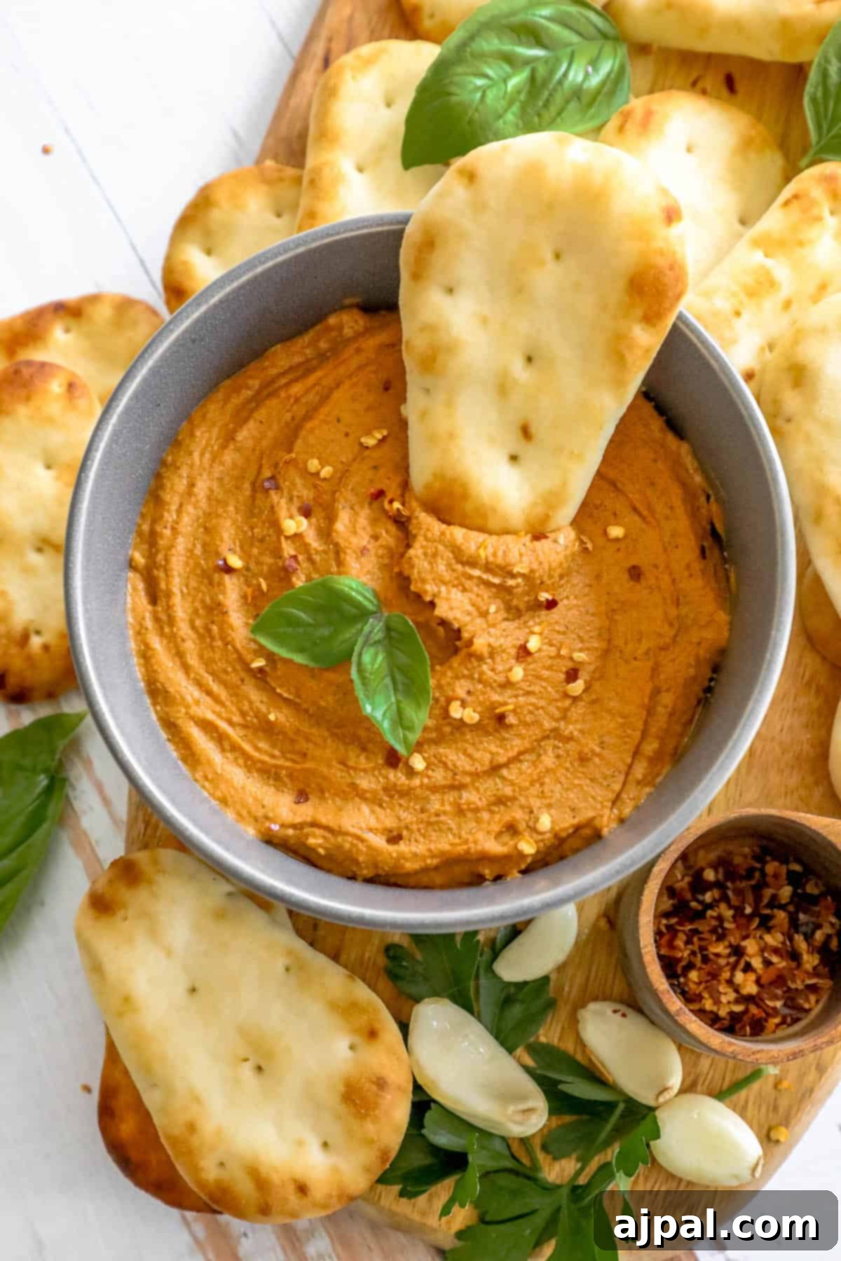 A ceramic bowl brimming with smooth, reddish hummus, adorned with a swirl of olive oil and a sprinkle of paprika, surrounded by an assortment of crisp crackers on a wooden surface.