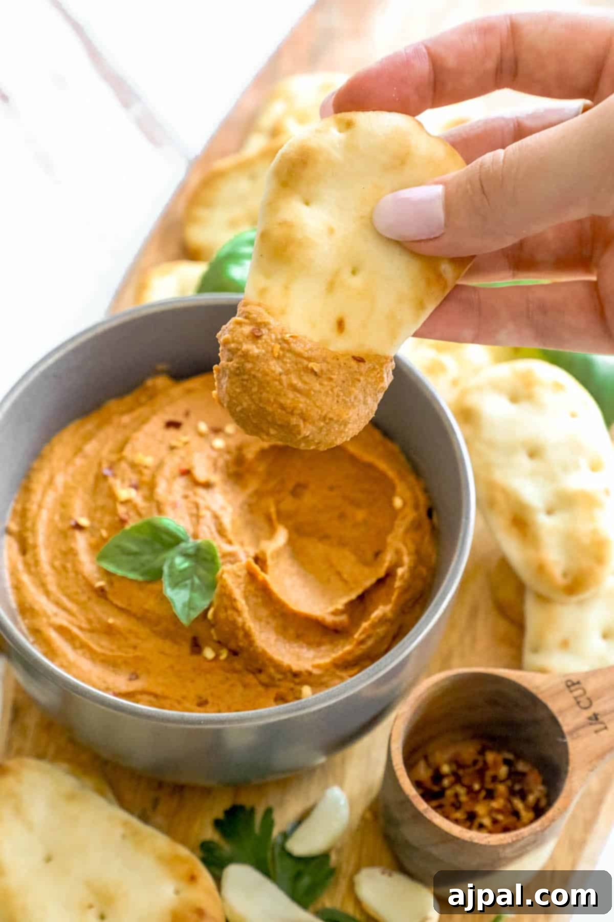 A hand reaches into a bowl of creamy pizza hummus with a crunchy pita chip, set against a backdrop of a wooden cutting board.
