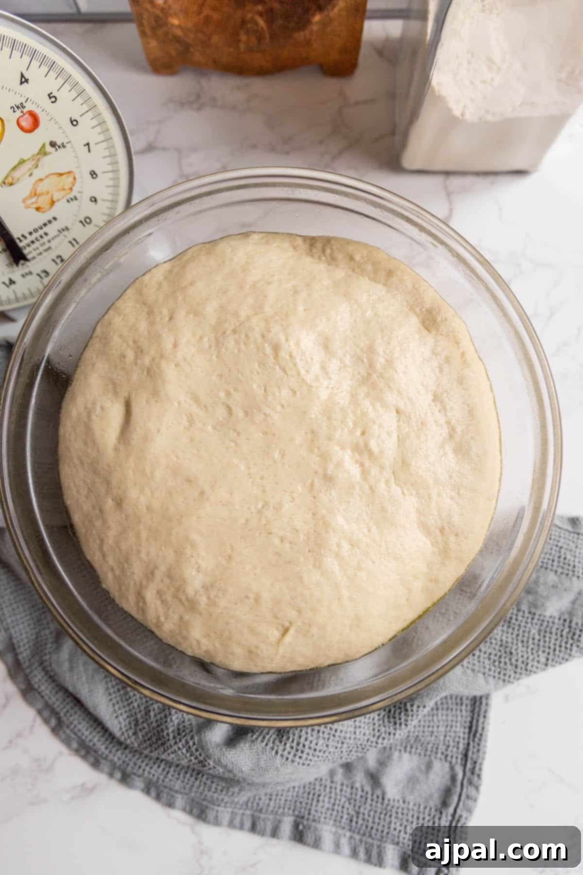 Pizza dough proofing in a clear glass bowl, next to a measuring cup, ready for preparation.