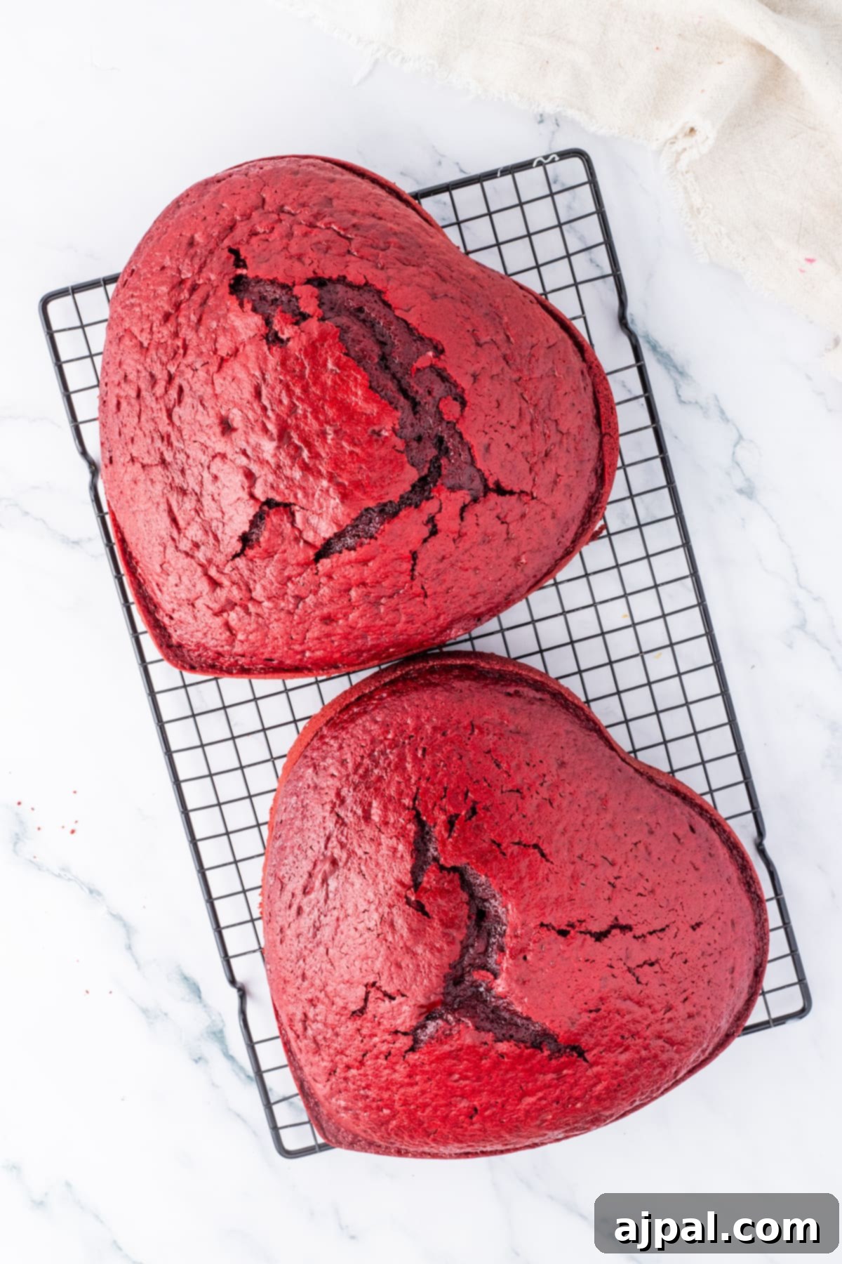 Two perfectly heart-shaped red velvet cakes rest elegantly on a cooling rack, awaiting their luscious frosting.