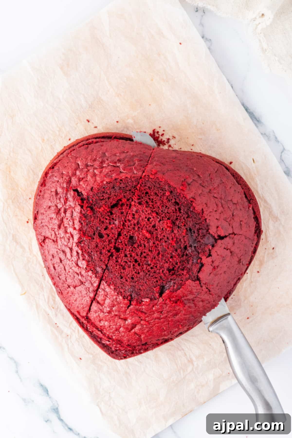 A heart-shaped Red Velvet Cake, beautifully frosted, sits with a knife placed gently on top, ready for slicing.