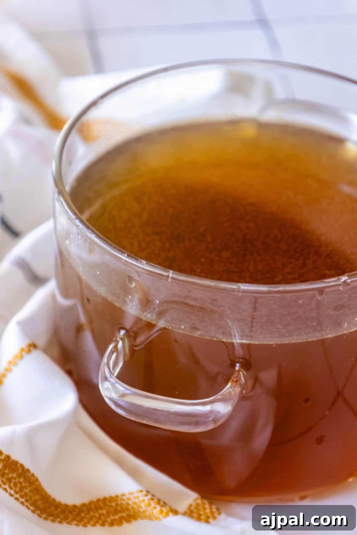 A glass bowl of golden chicken stock, steaming gently, resting on a white patterned towel, ready to be used.