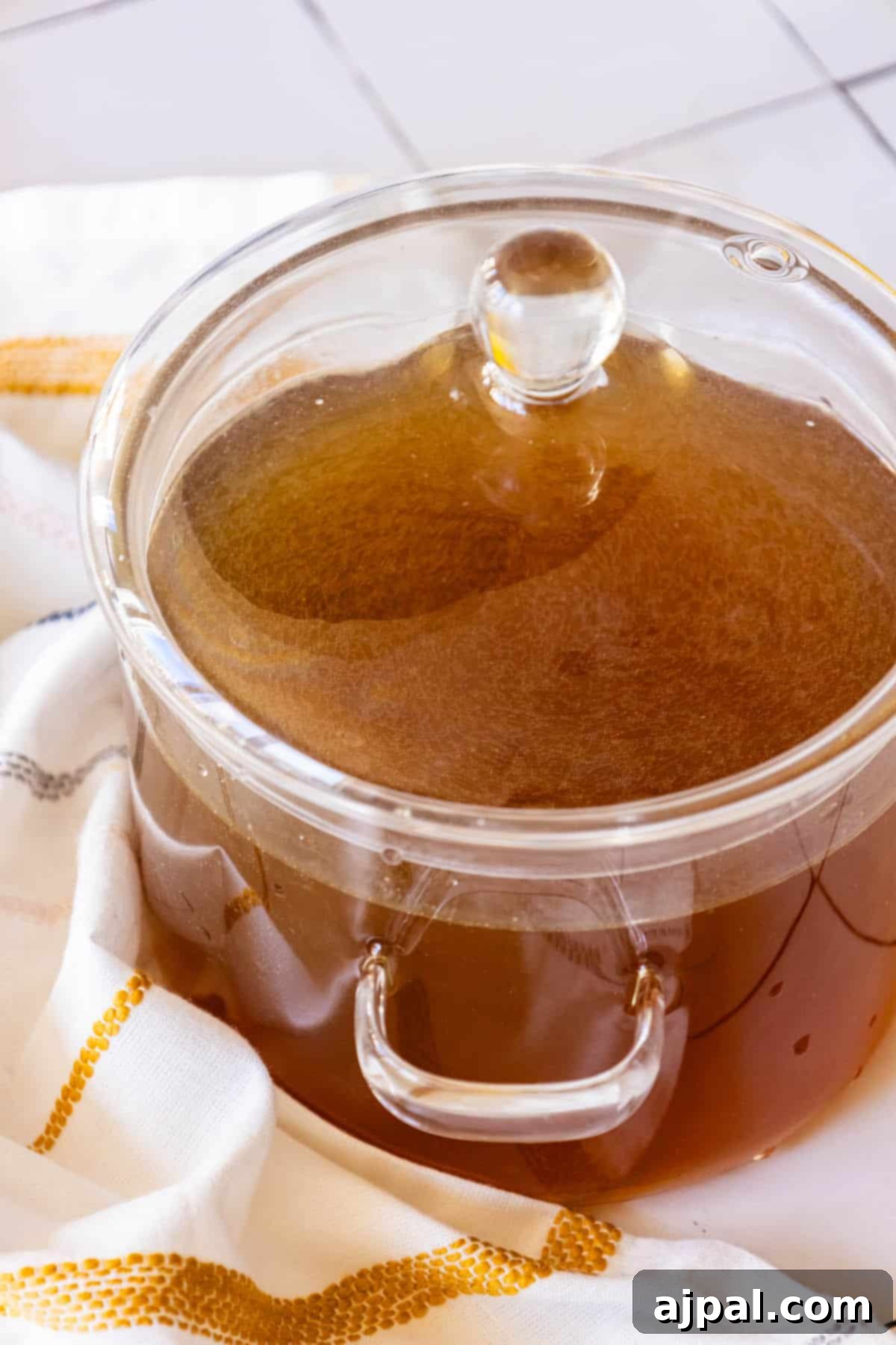 A glass pot filled with rich brown liquid, indicating freshly made chicken stock, sits on a pristine white towel.