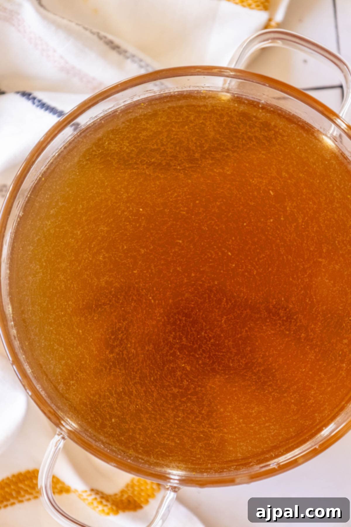 A beautiful glass bowl filled with golden homemade chicken stock, perfectly clear and ready for cooking, sits on a rustic table.