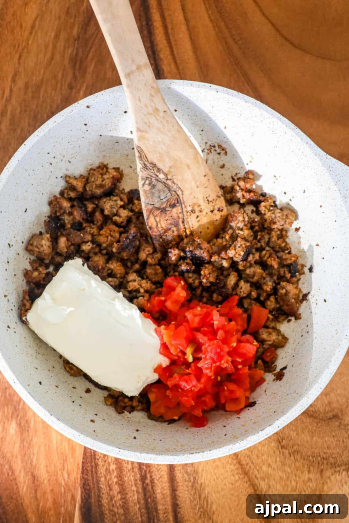A white bowl filled with cooked sausage, Rotel tomatoes, and softened cream cheese, ready to be mixed.