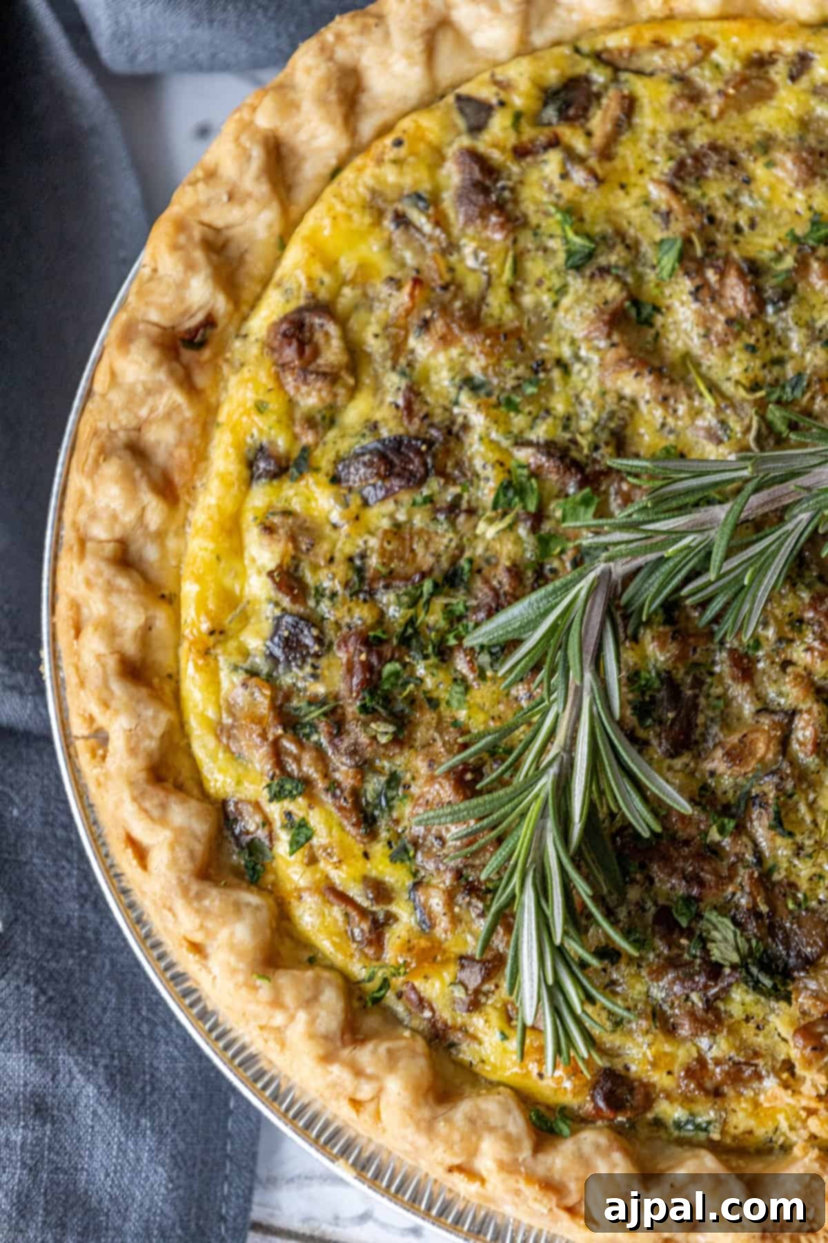 Forest Mushroom Quiche 7 A slice of mushroom quiche garnished with a sprig of rosemary on a white plate.