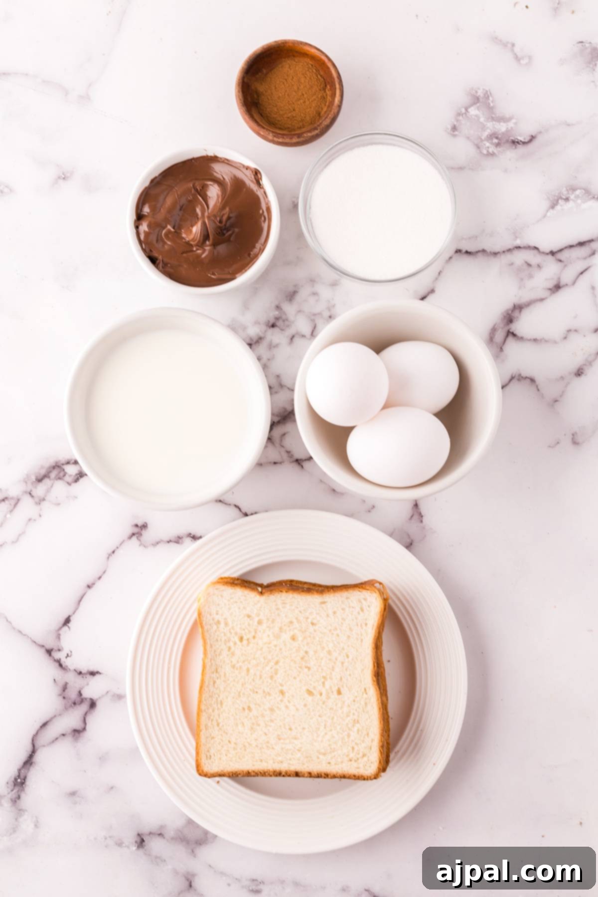 Ingredients for Nutella Stuffed French Toast Bites including bread, eggs, milk, and butter on a marble table.