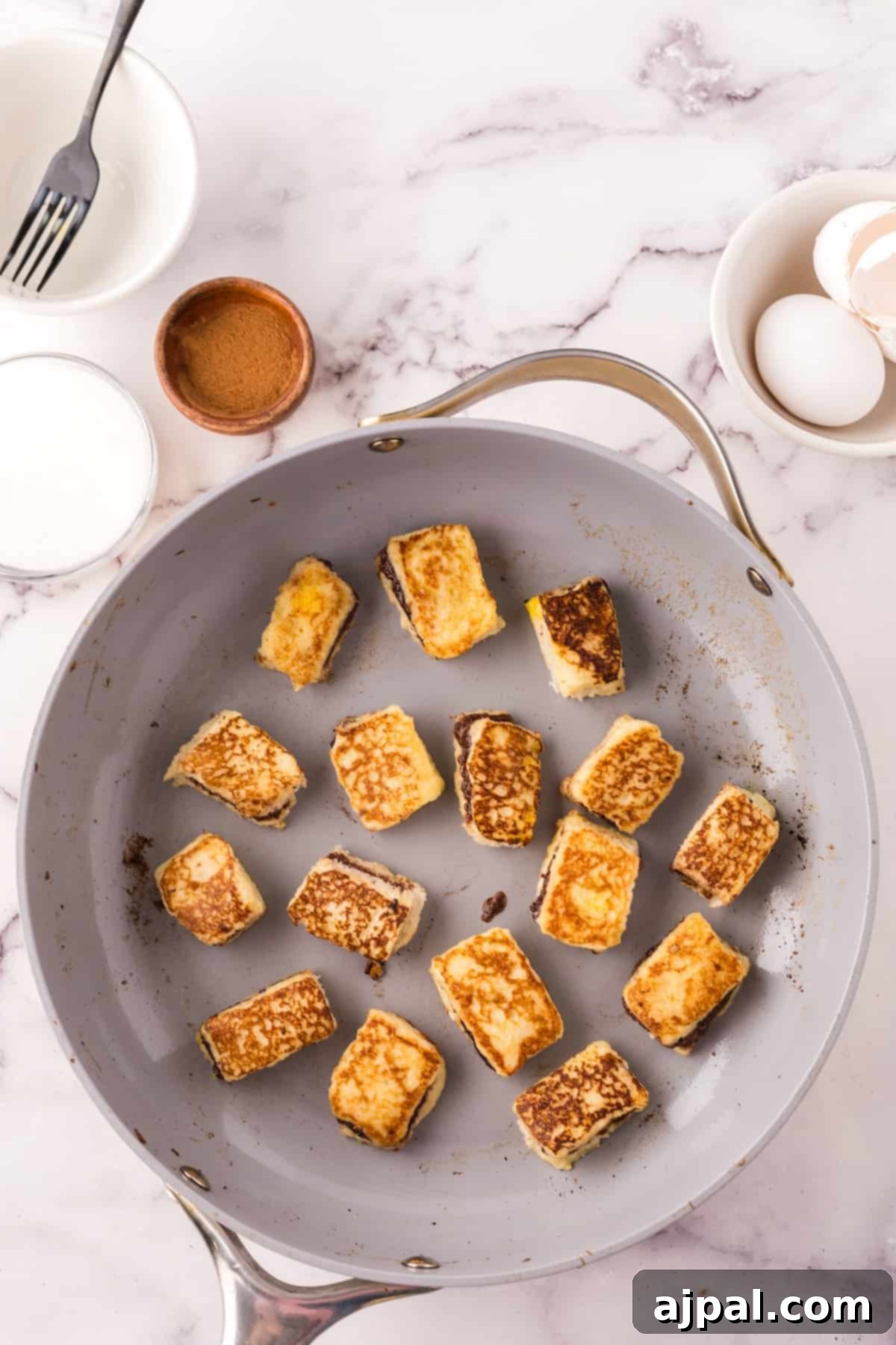 A non-stick pan with several Nutella Stuffed French Toast Bites cooking on a marble surface.