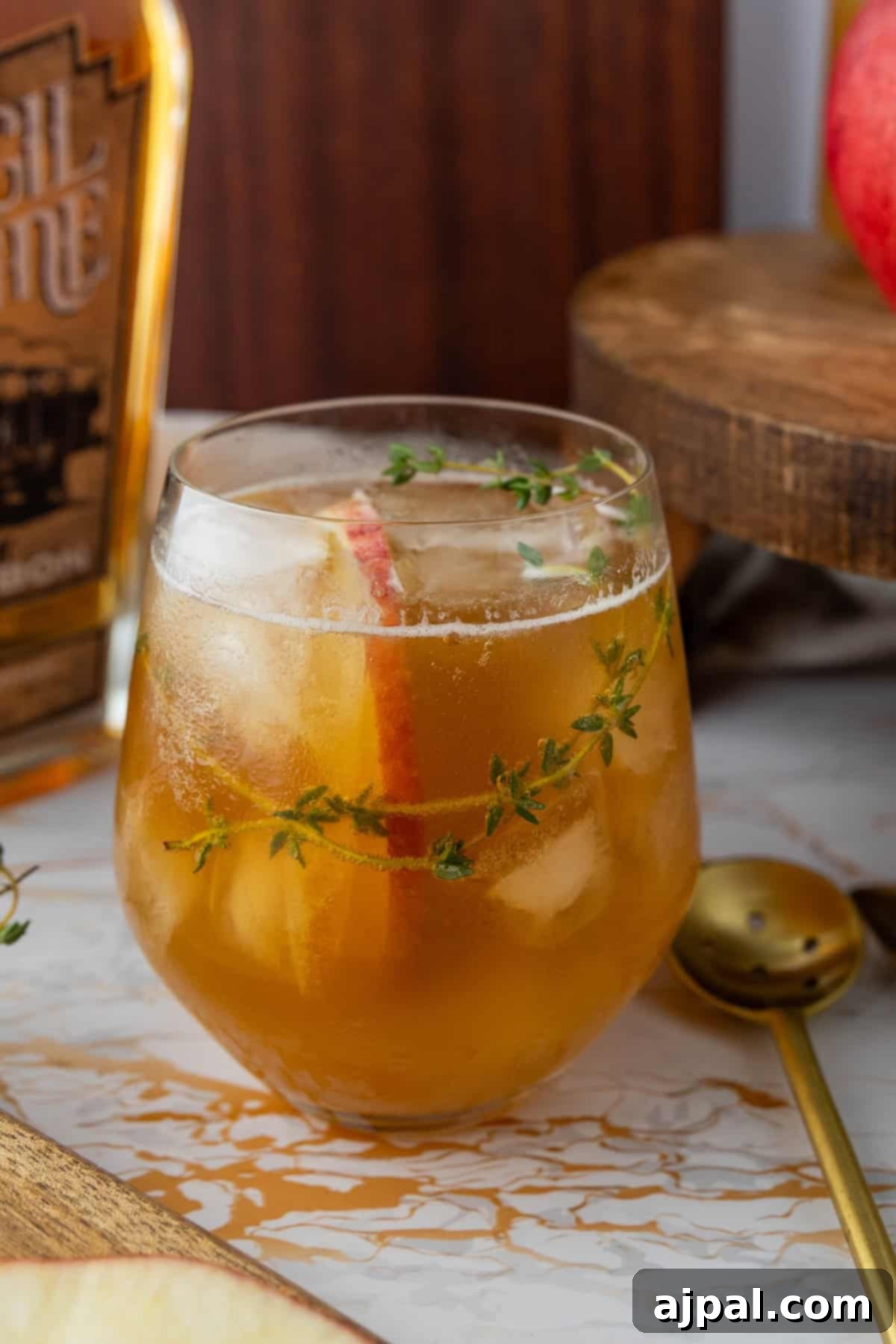 A glass of iced apple cocktail garnished with a sprig of thyme. A bottle and a spoon are visible in the background.