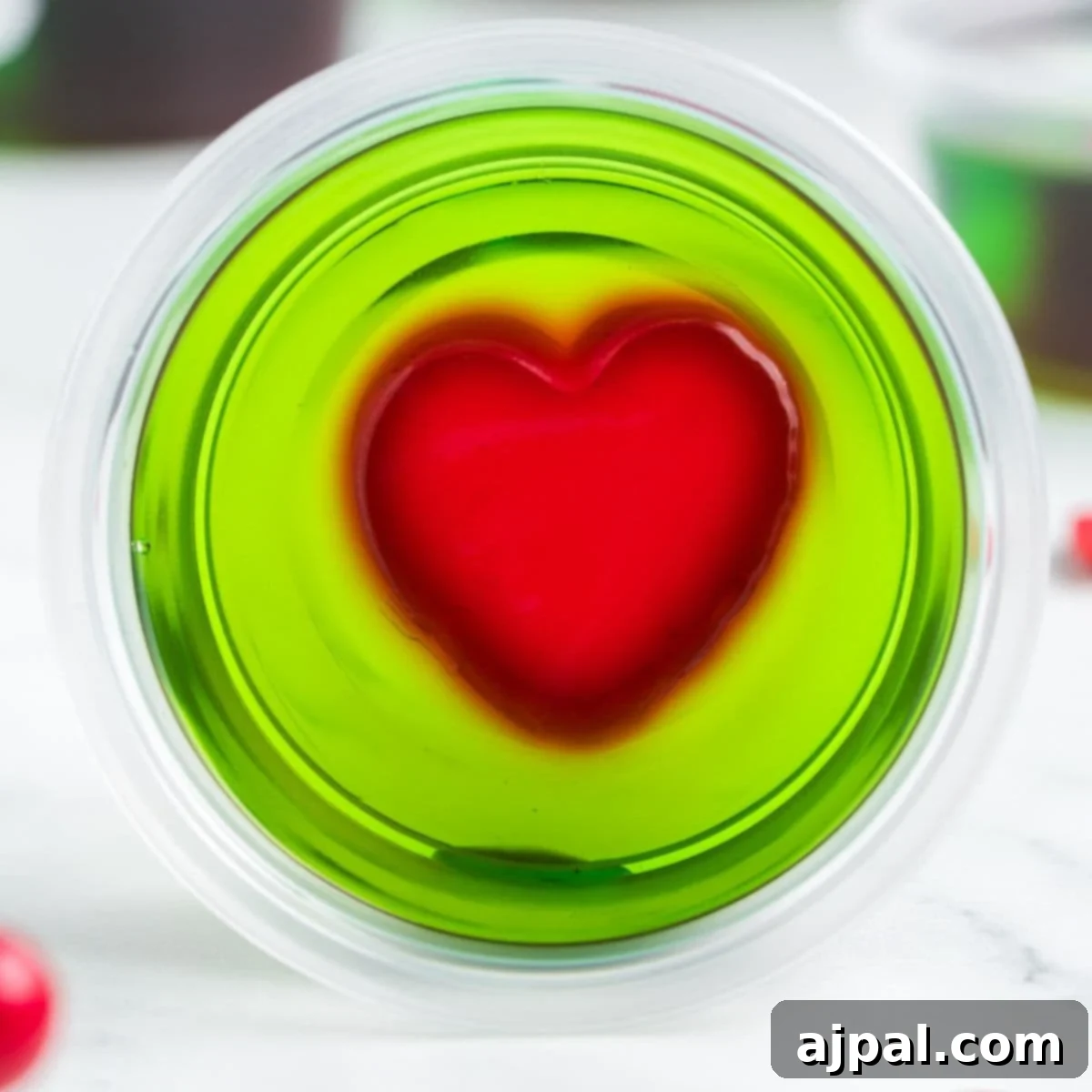 green jello shot with a red jello heart in the middle in a plastric cup on a table