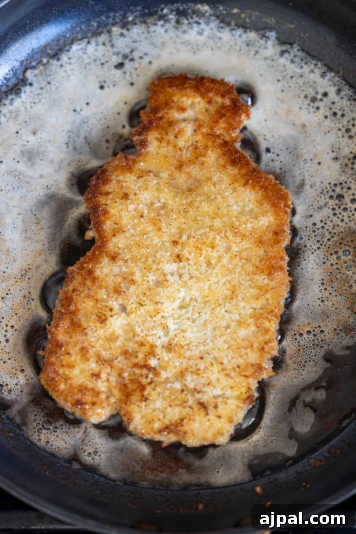 A breaded cutlet is being fried in a pan with bubbling oil and butter, revealing the golden brown crust typical of a classic Pork Schnitzel recipe.