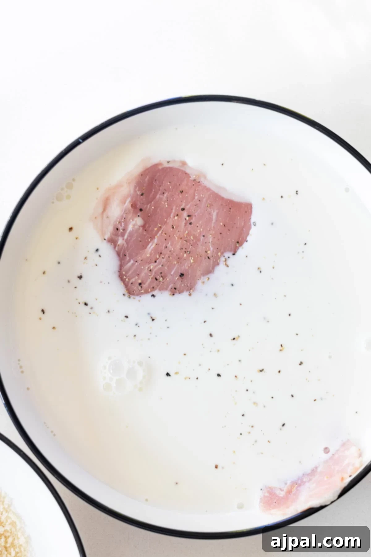 A raw pork chop soaking in a bowl of milk with black pepper sprinkled on top, perfect as the first step for a tender Pork Schnitzel recipe.