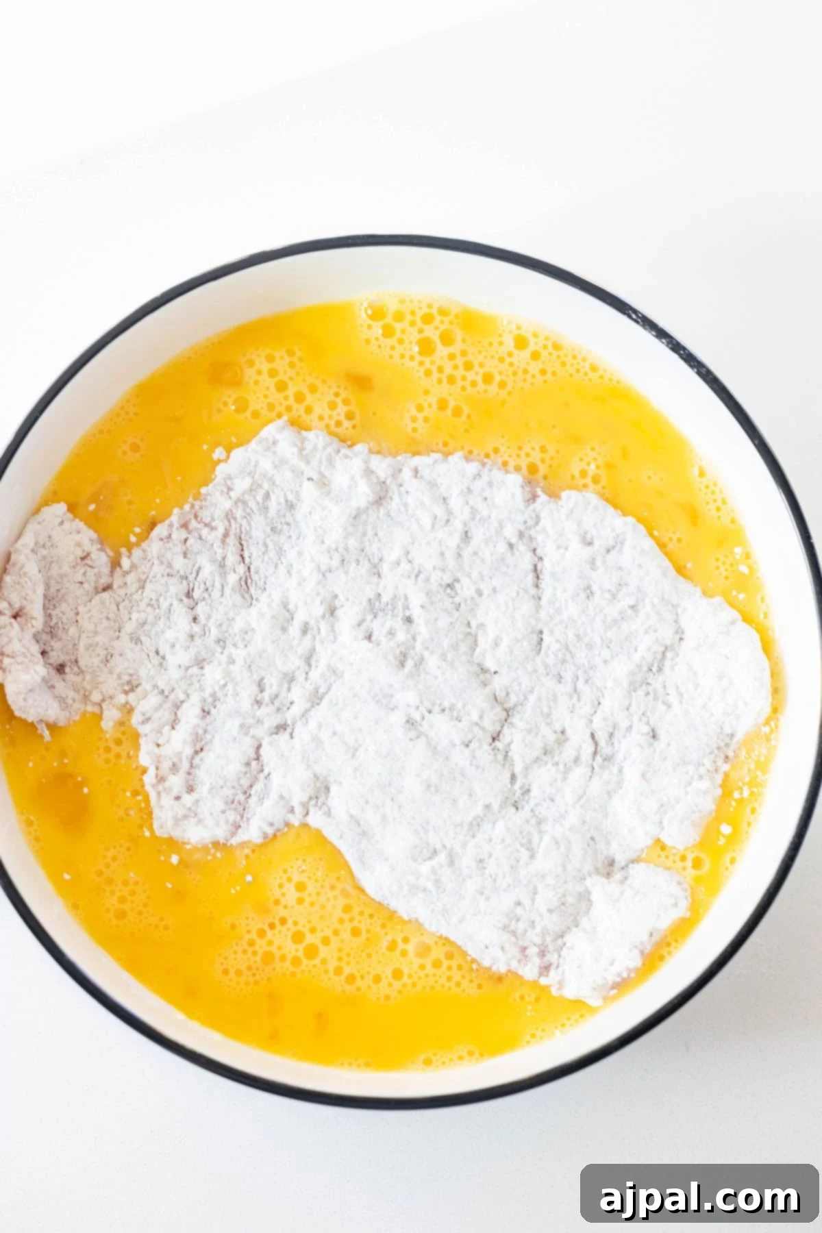 A bowl with beaten eggs and a piece of flour-coated meat being dipped into the mixture, perfect for preparing a classic Pork Schnitzel recipe, on a white surface.
