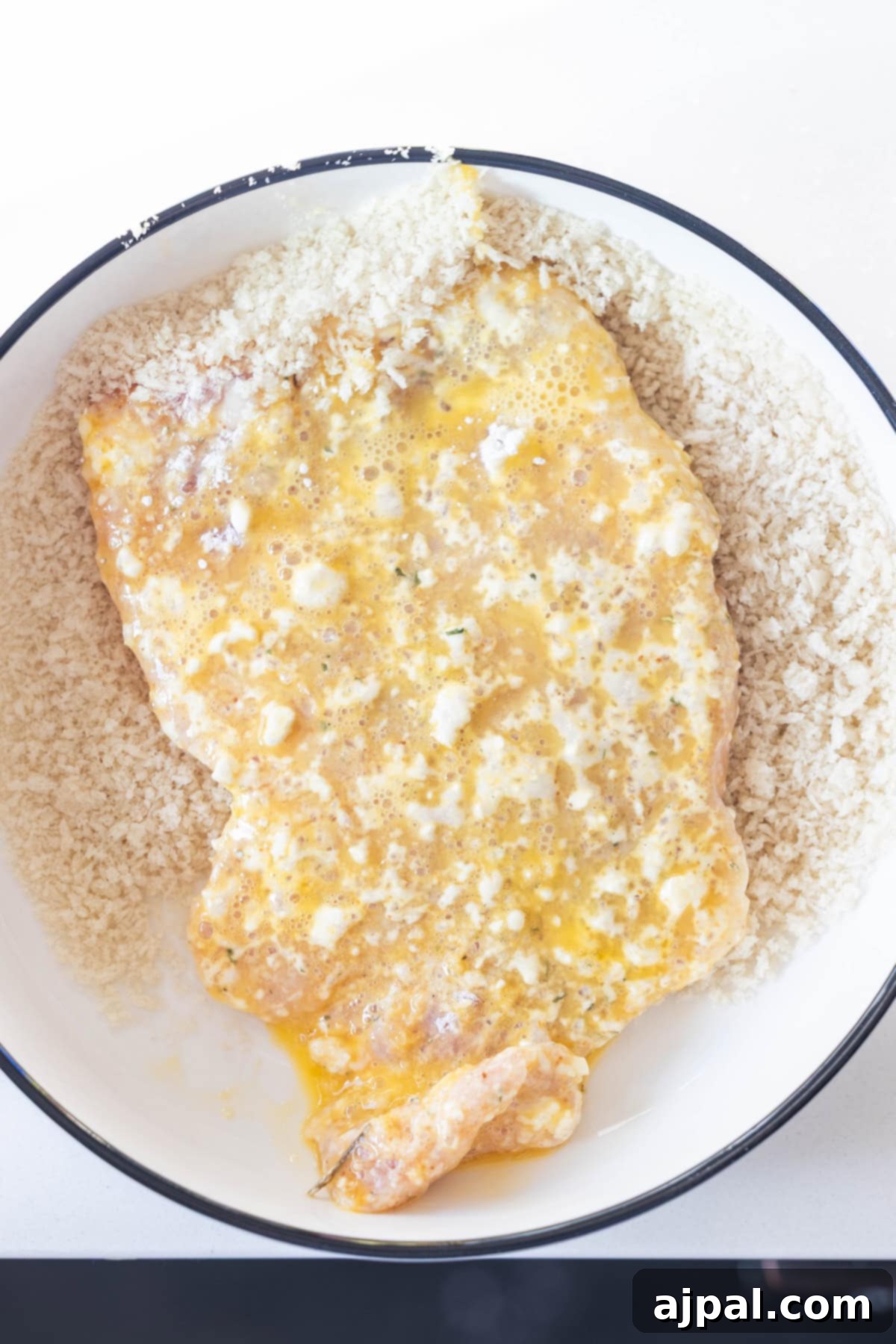 A raw breaded chicken cutlet coated in beaten egg is placed on a bed of panko breadcrumbs in a white shallow bowl, similar to the method used in a classic Schnitzel recipe.