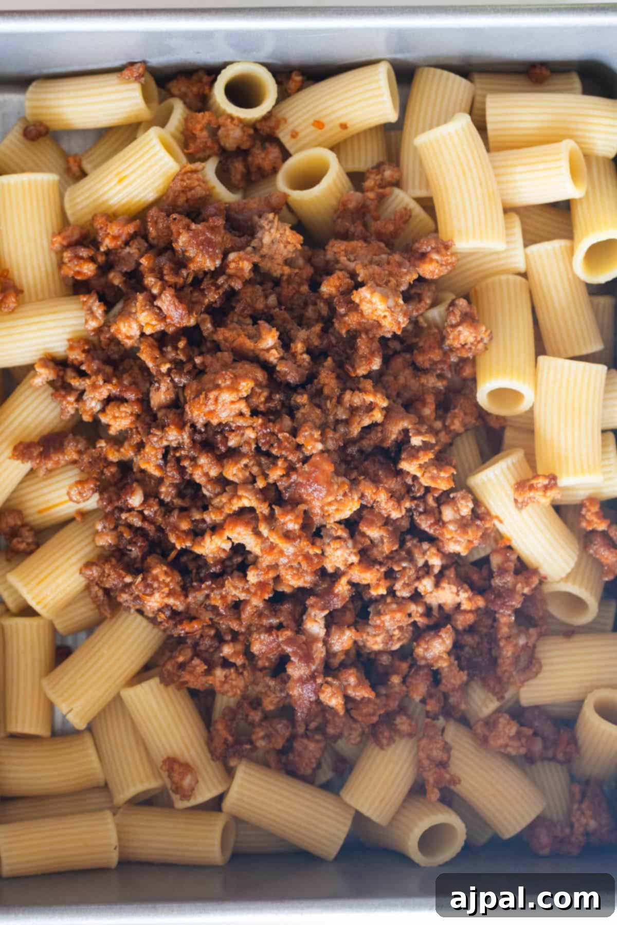 Cooked rigatoni pasta in a metal tray topped with crumbled ground meat in a red sauce.