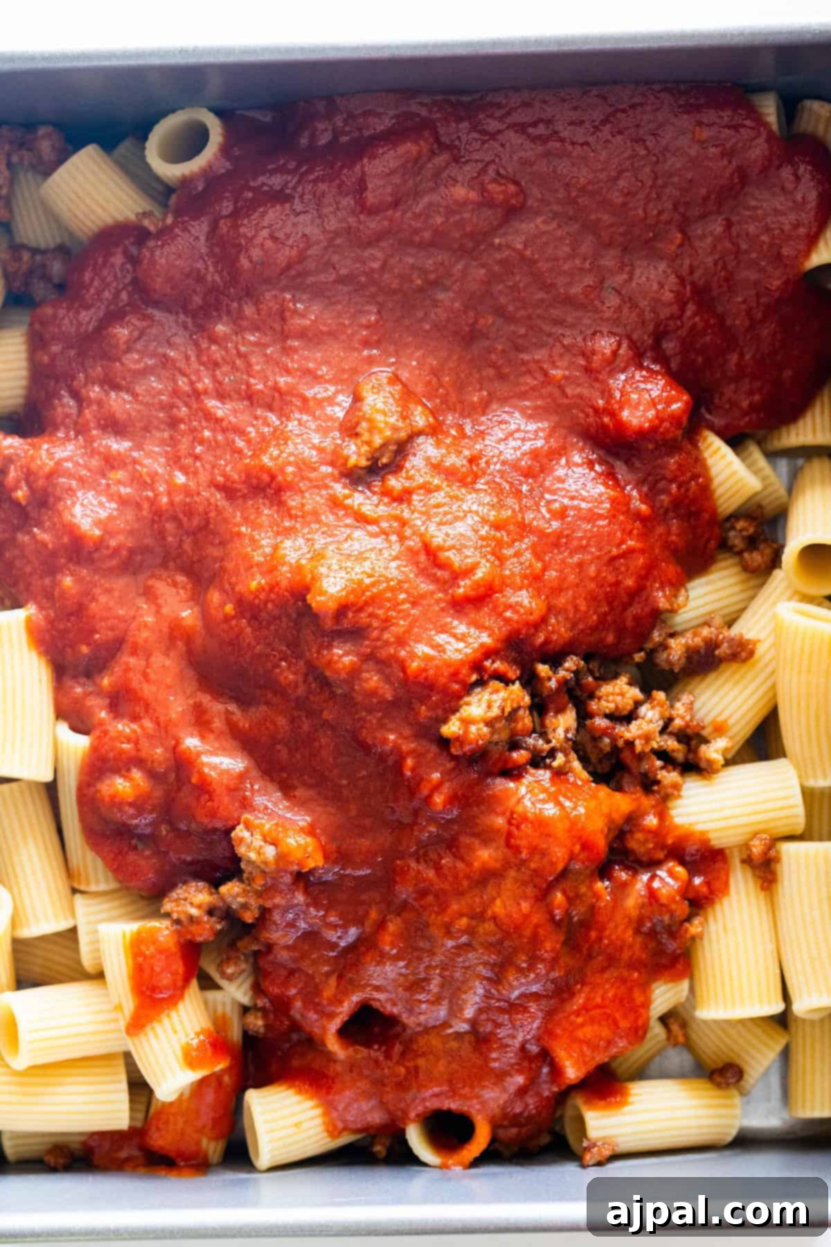 Cooked rigatoni pasta topped with ground meat and tomato sauce in a rectangular baking dish.
