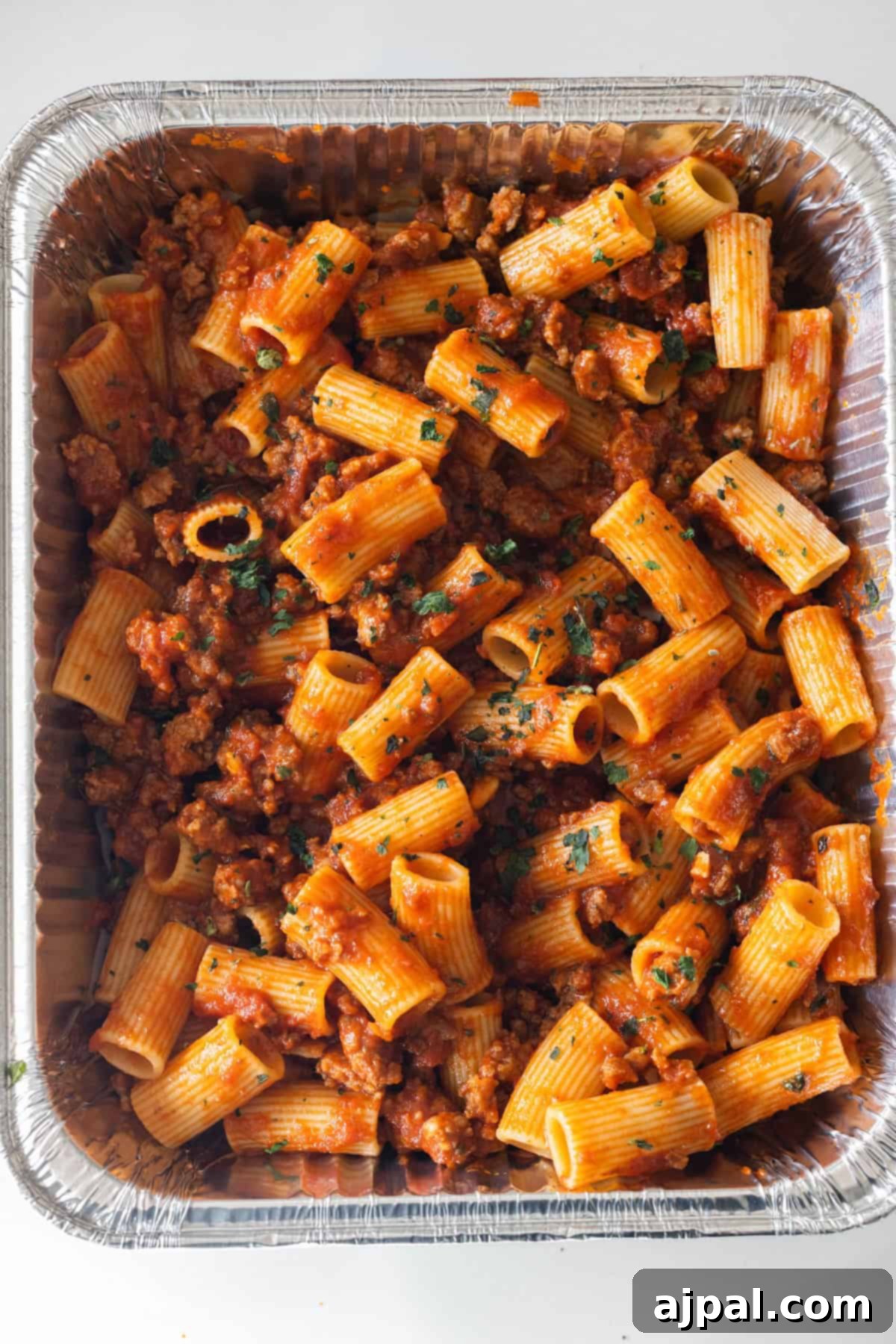 A large aluminum tray filled with cheesy baked rigatoni pasta, topped with ground meat and tomato sauce, and garnished with chopped herbs.