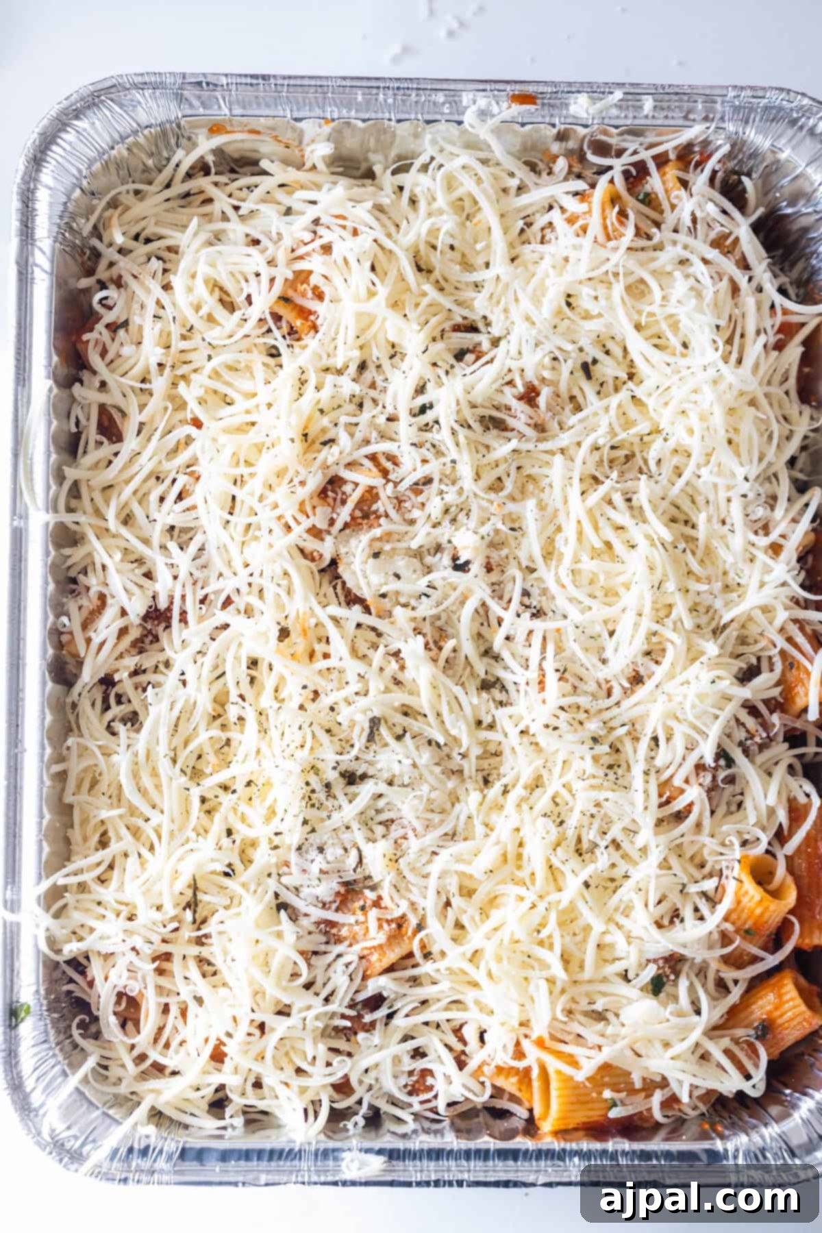 A foil tray filled with cheesy baked rigatoni pasta, topped with shredded cheese and herbs, ready to be baked.