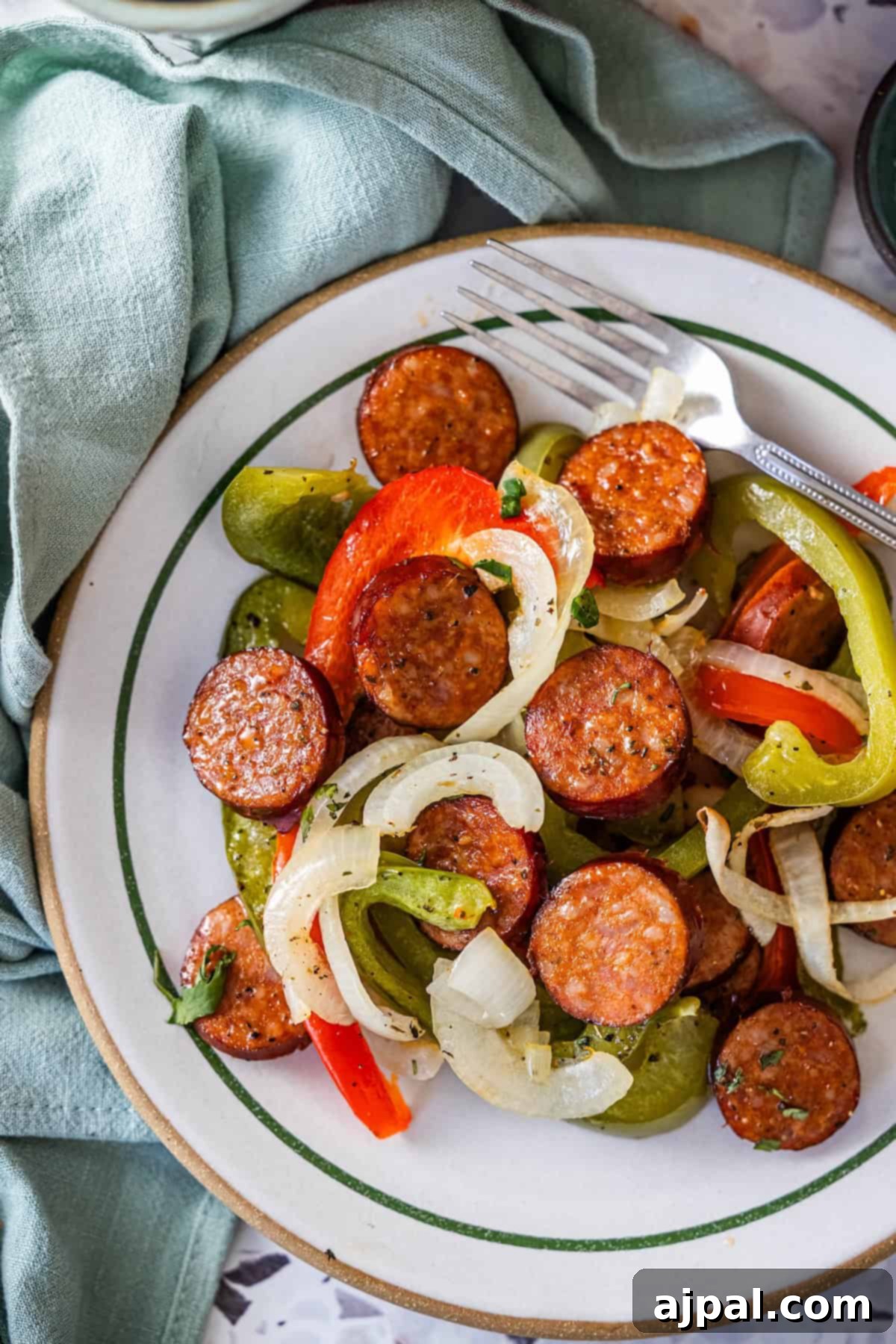 A plate of Sheetpan Kielbasa with sautéed onions, and red and green bell peppers, served alongside a fork and knife.
