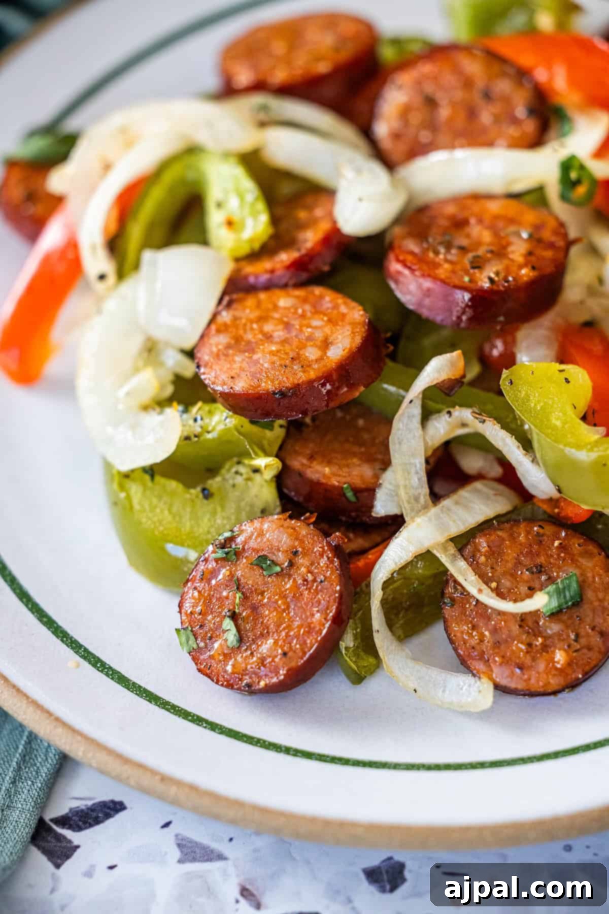 A plate of sheetpan Kielbasa, cooked onions, and vibrant peppers arranged on a white plate with a green rim.