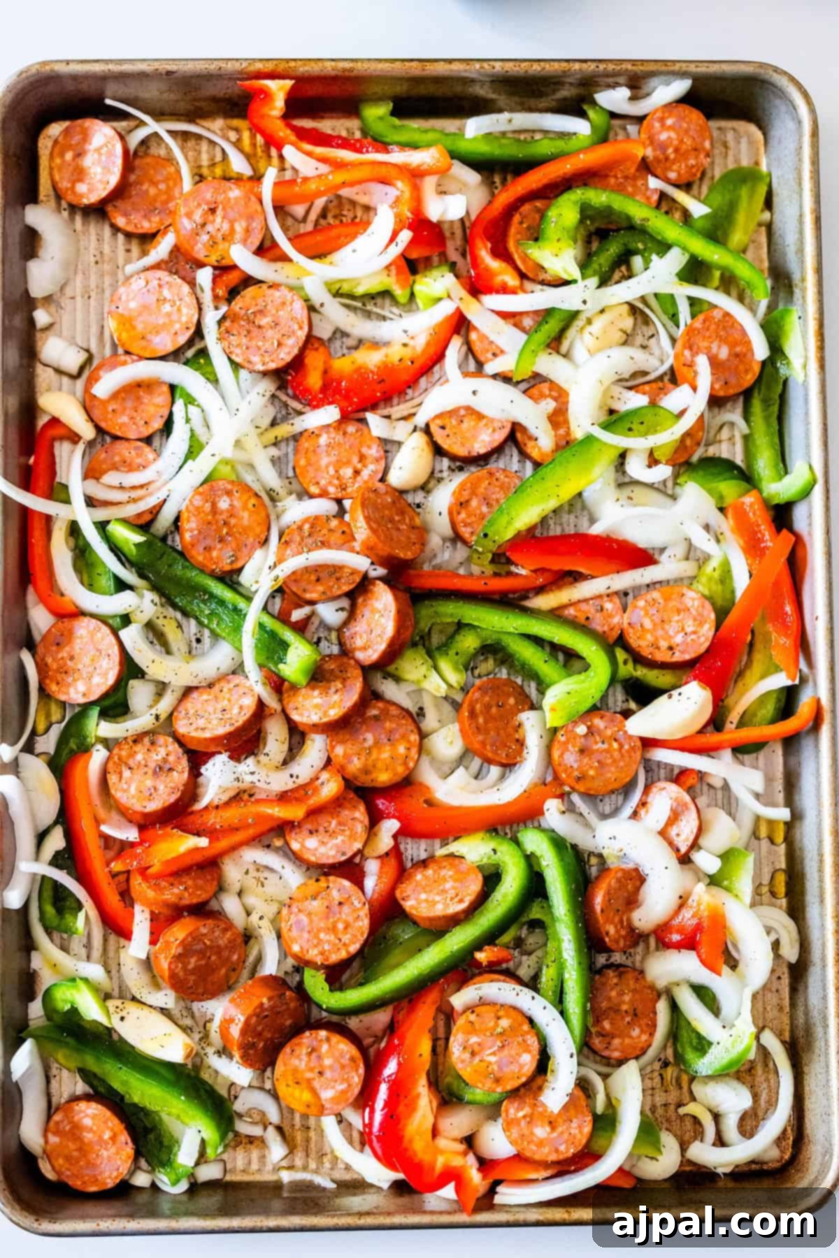 Sliced kielbasa, green and red bell peppers, and onions spread on a sheetpan lined with parchment paper, ready to be baked.