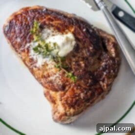 A cooked sirloin steak topped with a dollop of butter and a sprig of thyme on a white plate with a green rim, with a fork and knife beside it.