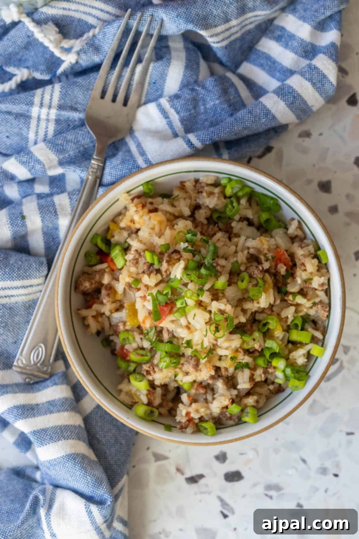 A bowl of rice mixed with ground meat, chopped vegetables, and topped with sliced green onions sits next to a fork on a blue and white striped cloth.