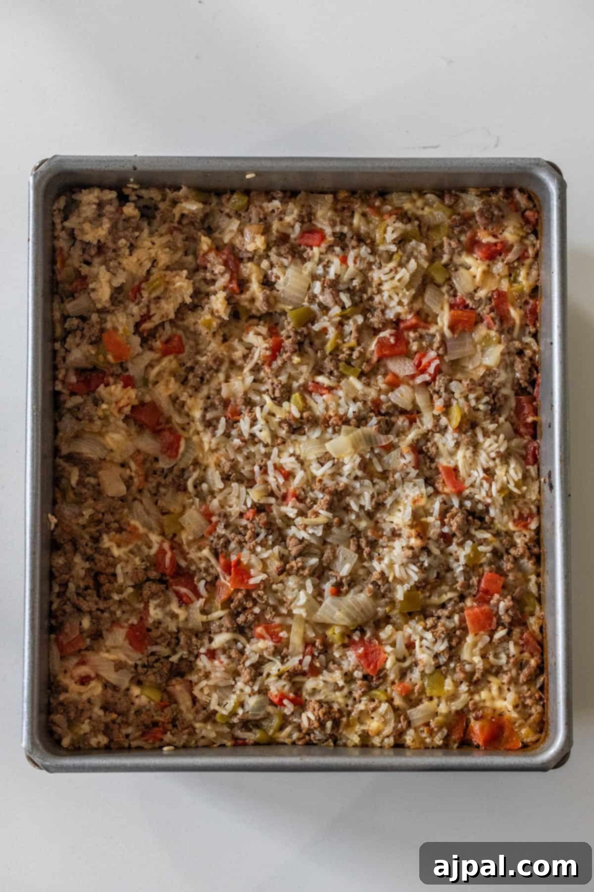 A square baking pan filled with Baked Dirty Rice casserole, featuring ground meat, rice, chopped onions, red bell peppers, and green bell peppers.