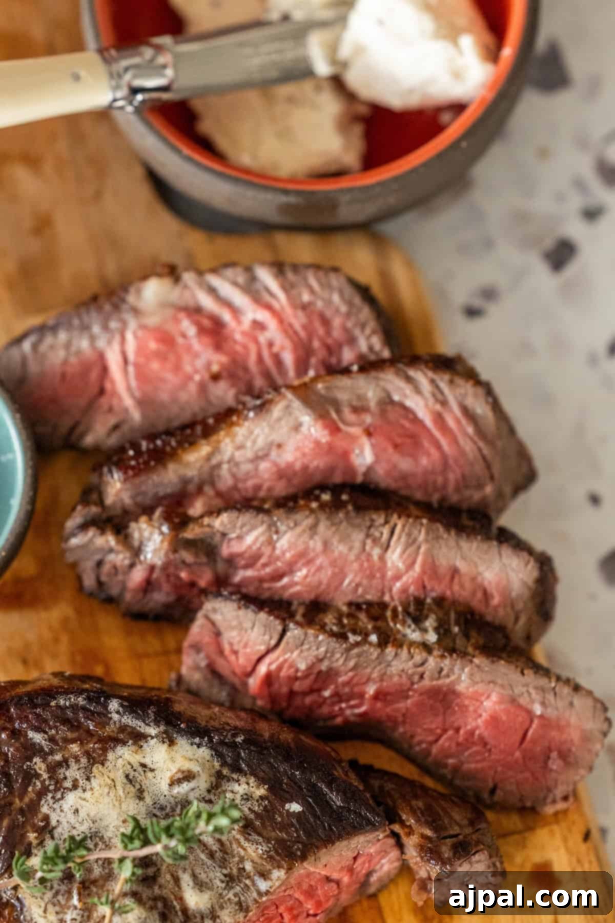 Slices of perfectly cooked, medium-rare sirloin steak elegantly arranged on a wooden board, accompanied by a sharp knife and a bowl of creamy, decadent garlic herb sauce in the soft-focus background.