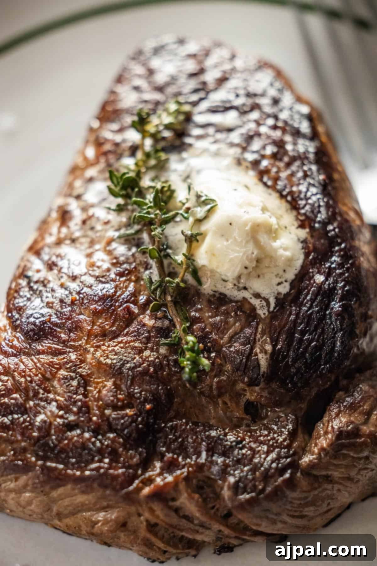 An enticing close-up shot of a flawlessly cooked Garlic Herb Sirloin, crowned with a luscious pat of melting butter and a delicate sprig of fresh thyme, presented on a pristine white plate.