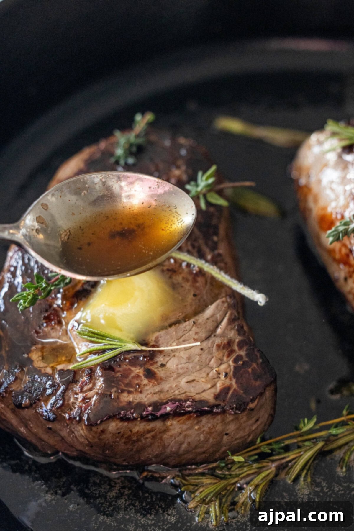 A spoon delicately drizzles melted butter and fragrant pan juices over a perfectly seared sirloin steak, garnished with fresh rosemary and thyme, all within the confines of a black skillet. A culinary masterpiece in progress.