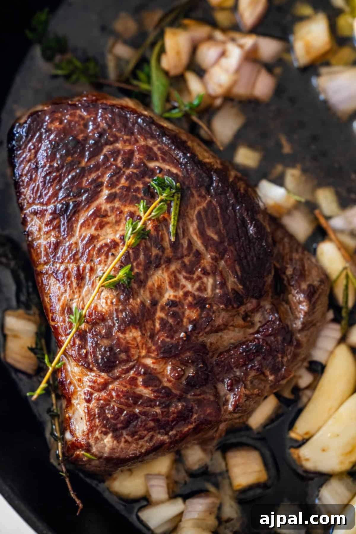 A seared sirloin steak nestled in a pan, surrounded by caramelized chopped onions, golden garlic cloves, and aromatic sprigs of fresh herbs. A symphony of flavors waiting to be enjoyed.