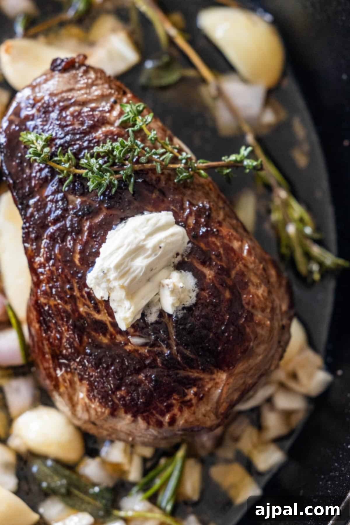 A perfectly cooked sirloin steak, generously topped with a dollop of melting butter and a sprig of fresh thyme, surrounded by aromatic garlic herb cloves and fresh herbs in a pan. A feast for the senses.