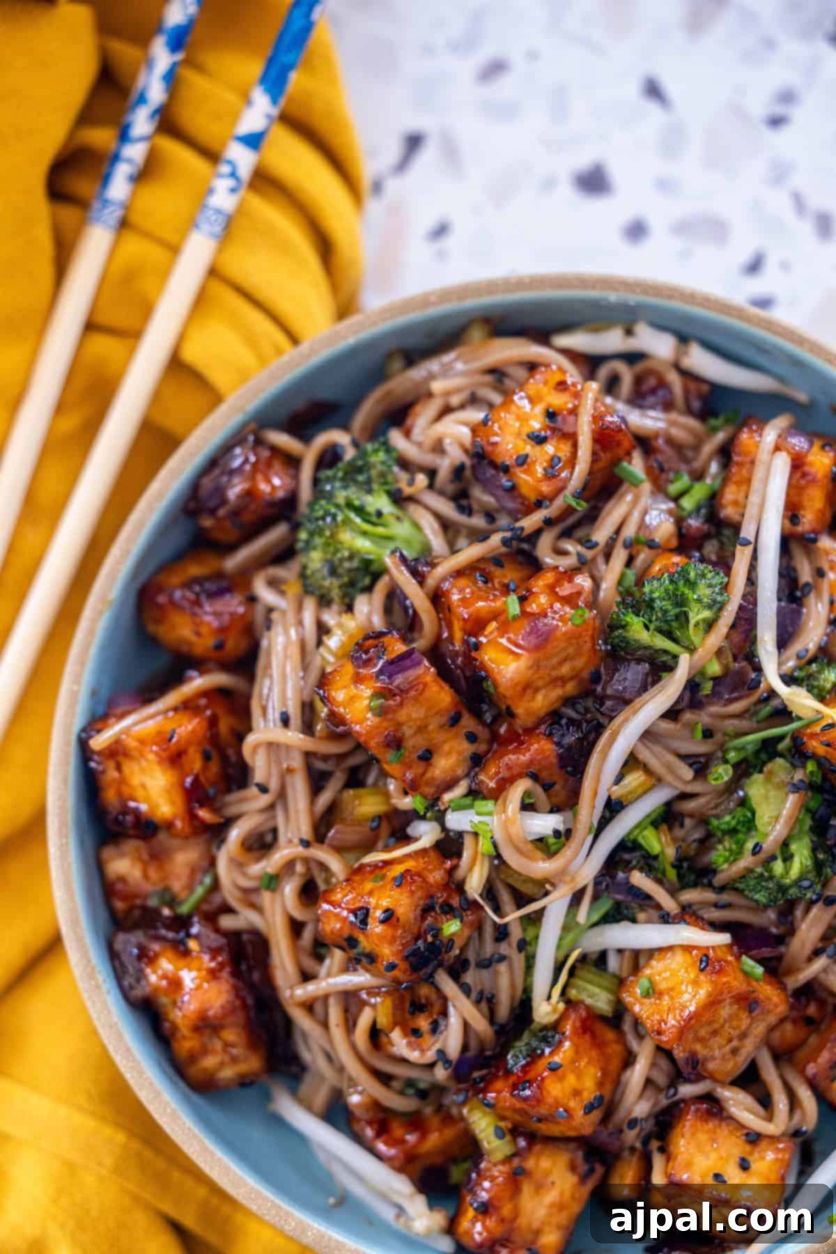 A bowl of crispy tofu and soba noodle stir-fry next to chopsticks on a yellow surface.