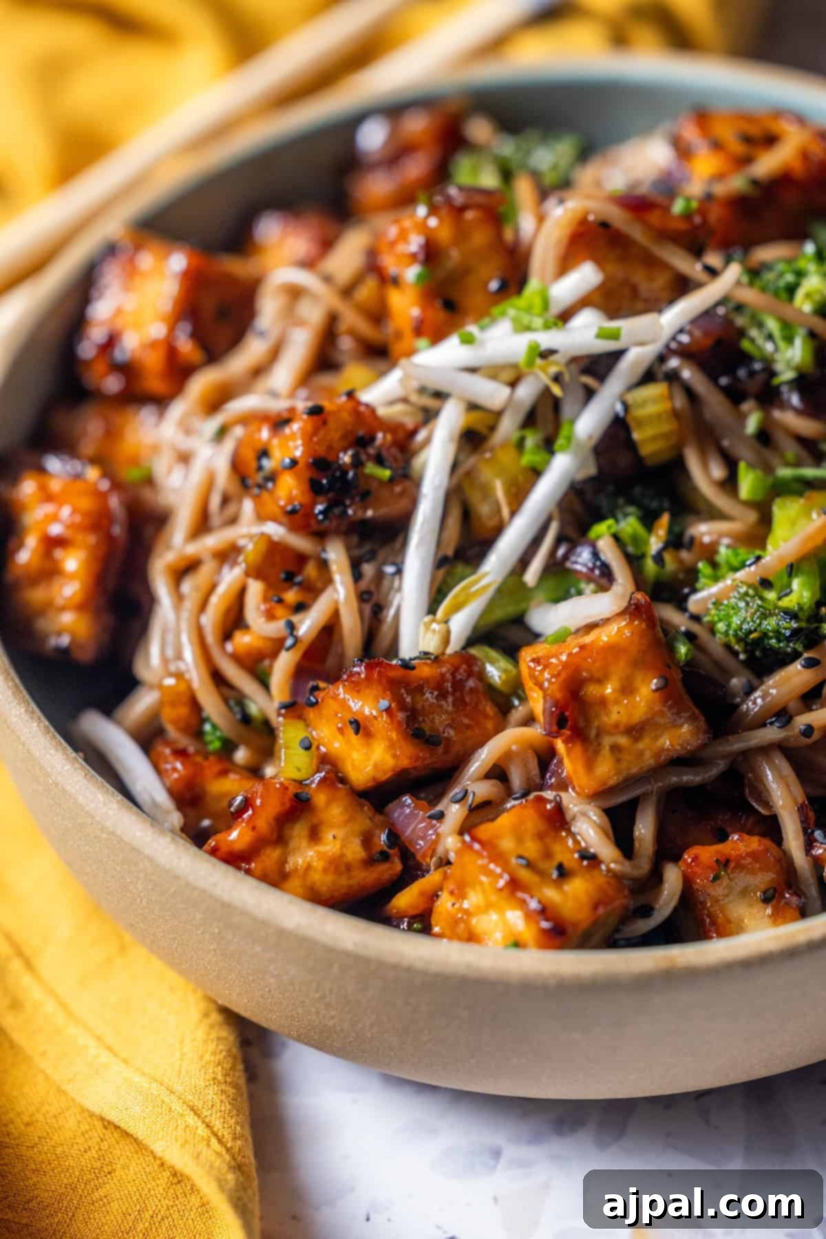 A bowl of stir-fried soba noodles with crispy tofu, broccoli, bean sprouts, and a rich, glossy sauce, garnished with chopped chives and sesame seeds.