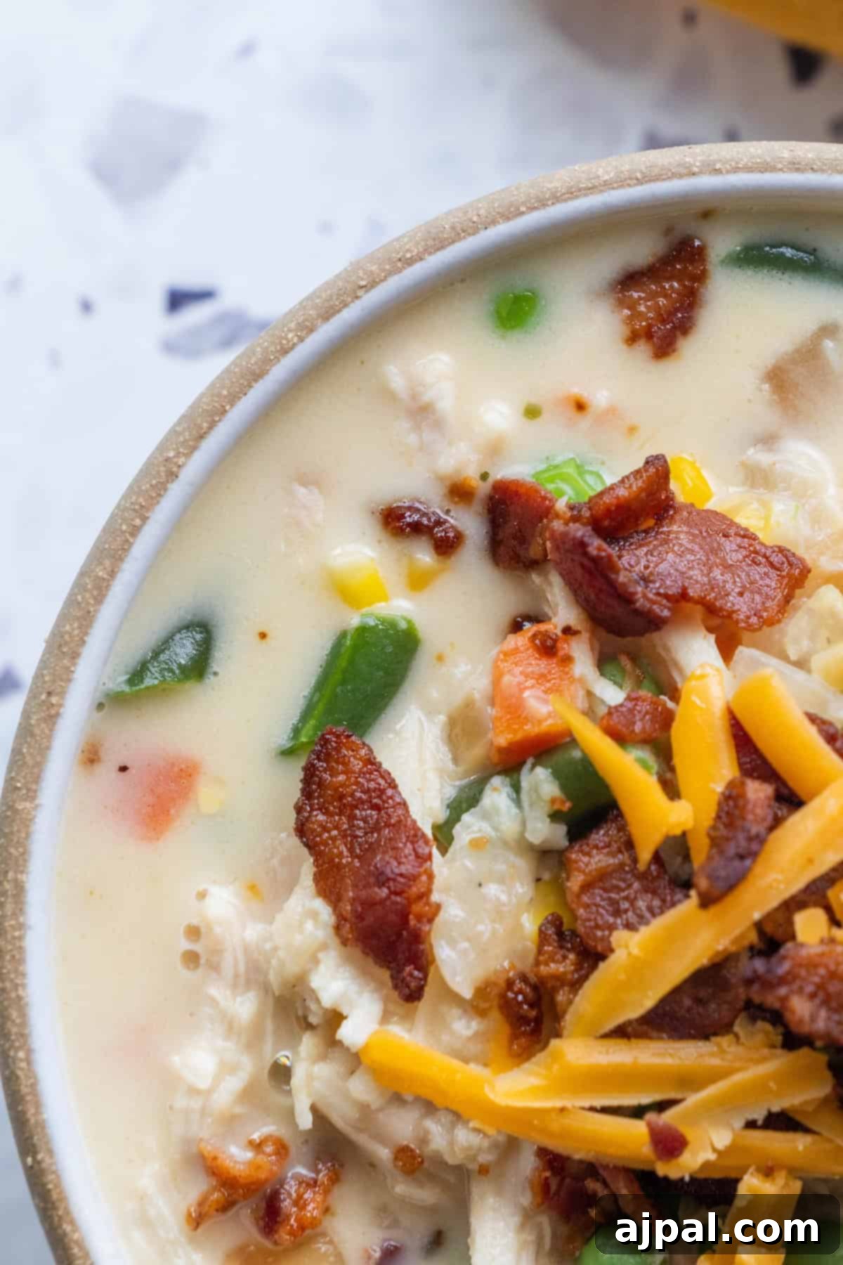A close-up of creamy chicken and corn chowder topped with crispy bacon pieces, shredded cheddar cheese, and chopped vegetables in a beige bowl.