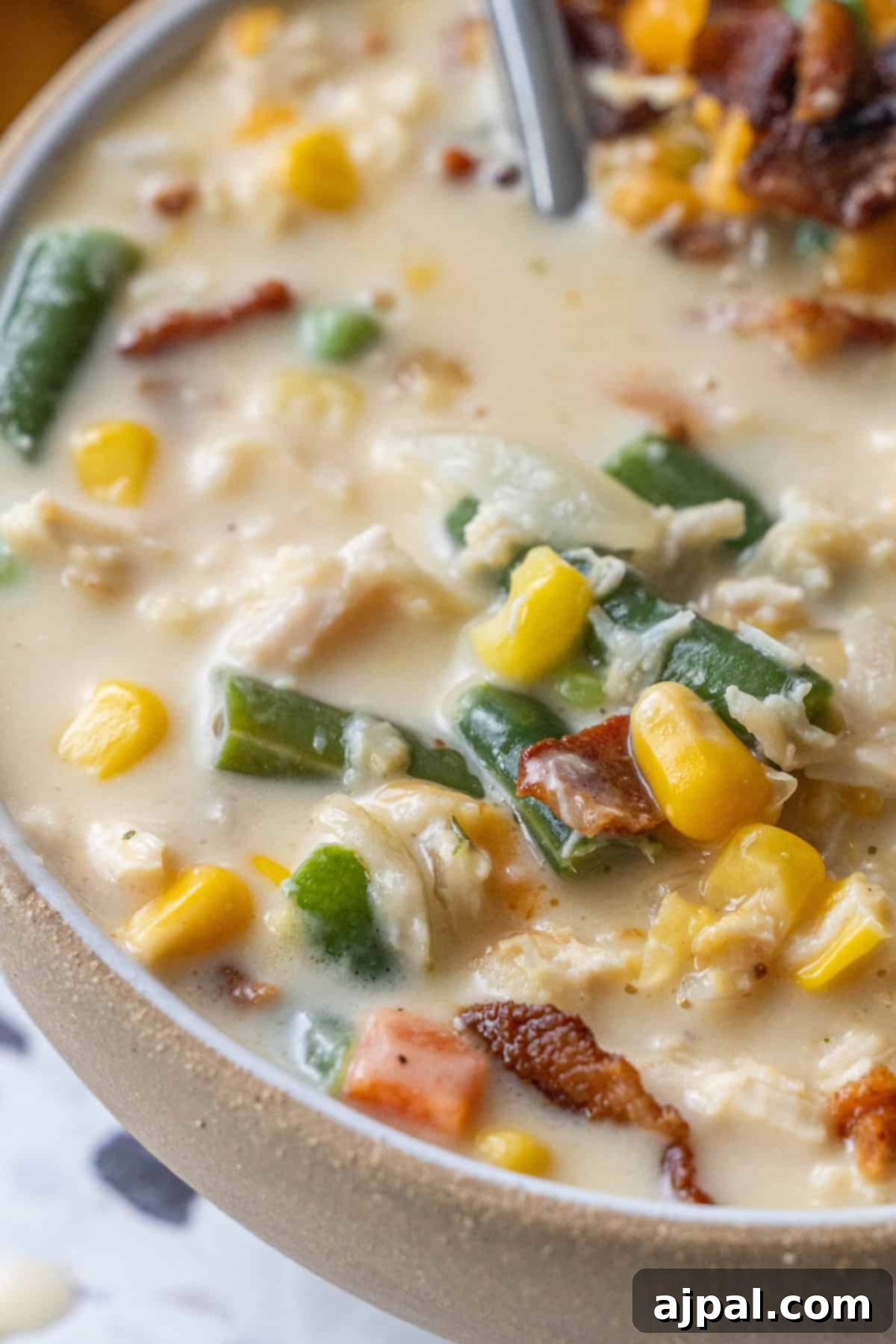 A close-up of a creamy chicken corn chowder with green beans, shredded chicken, and pieces of bacon in a beige bowl.