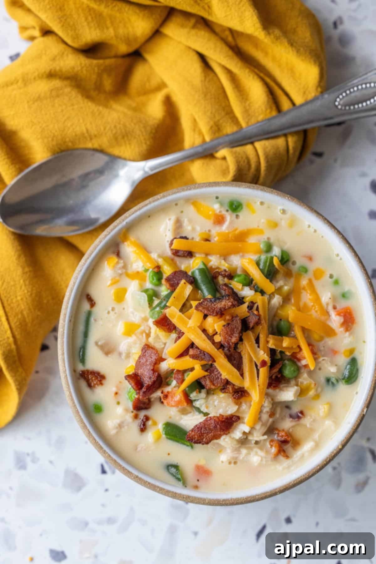 A bowl of creamy corn chowder with mixed vegetables, shredded cheddar cheese, and crispy bacon bits, placed on a marble surface next to a spoon and a yellow cloth.
