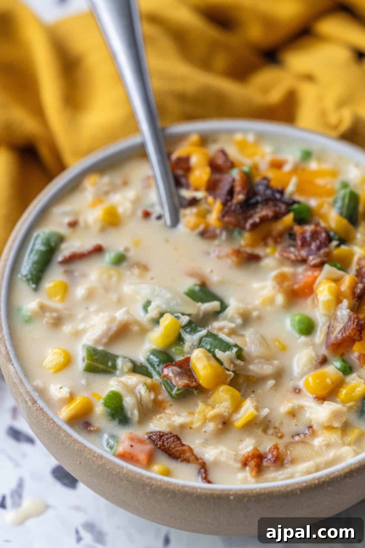 A bowl of creamy corn chowder featuring green beans, carrots, peas, shredded chicken, and crispy bacon pieces, with a spoon inside the bowl.