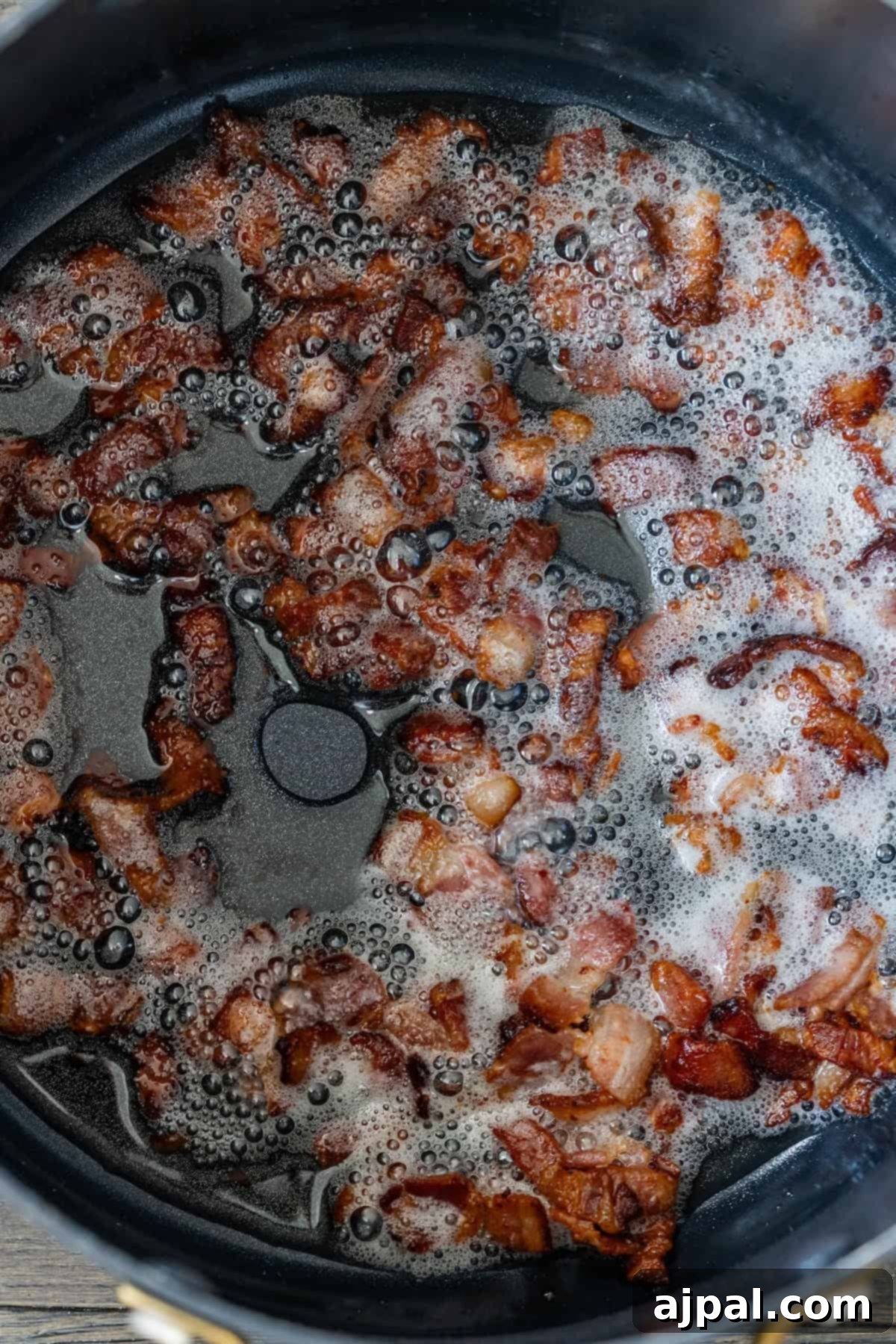 Crispy bacon pieces cooking in a black pan with bubbling grease and foam—perfect for topping cheesy corn chowder.