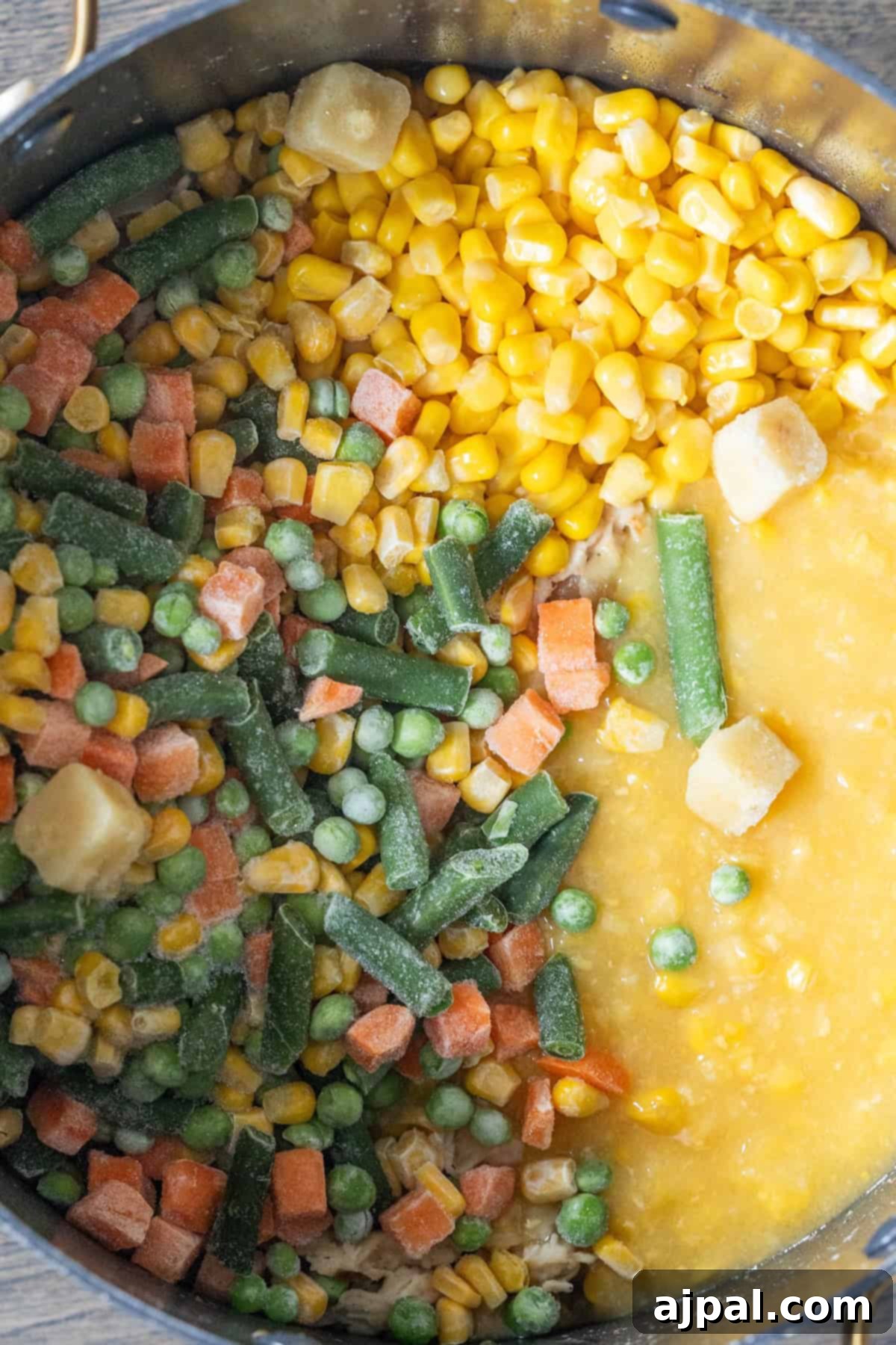 A pot contains frozen mixed vegetables, frozen corn, and a thick yellow liquid, partially blended together for a hearty Cheesy Corn Chowder.