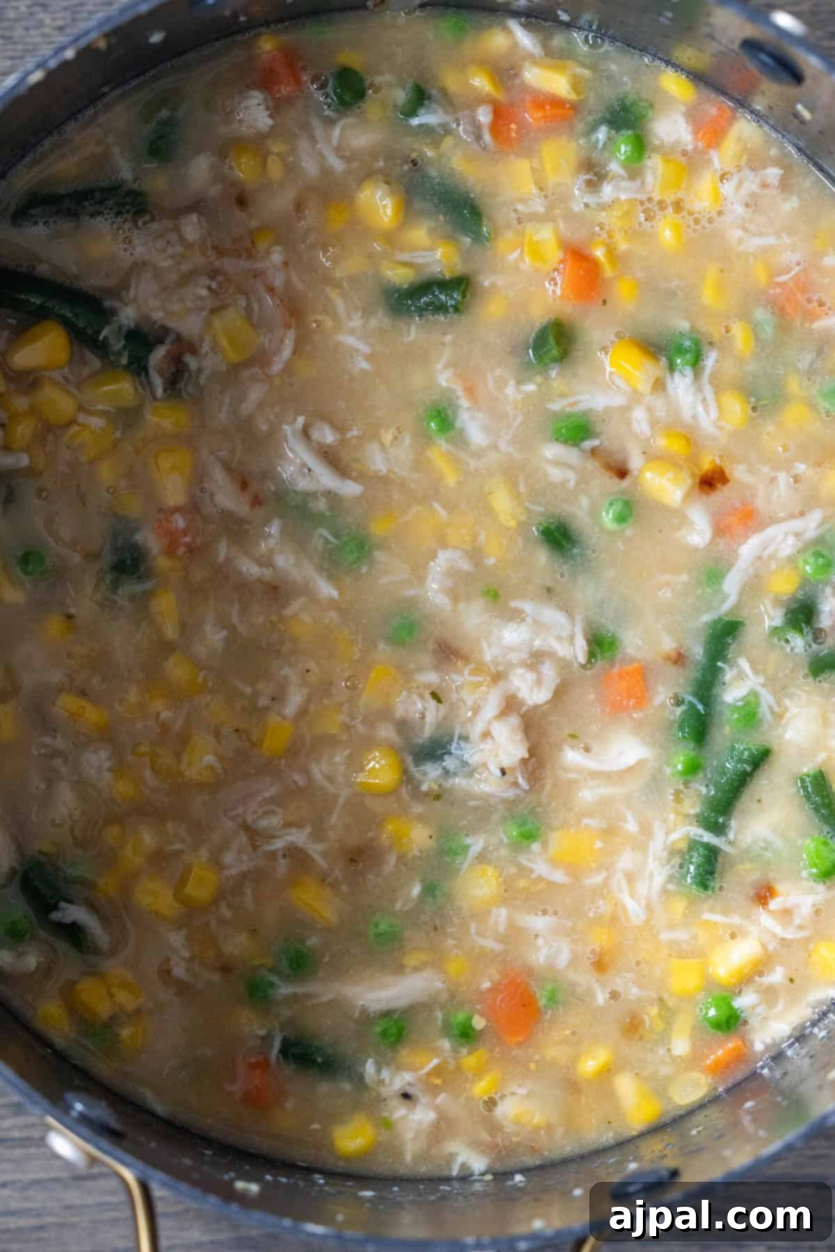 A close-up of a pot filled with thick cheesy corn chowder featuring bacon, green beans, peas, carrots, and tender shredded chicken.
