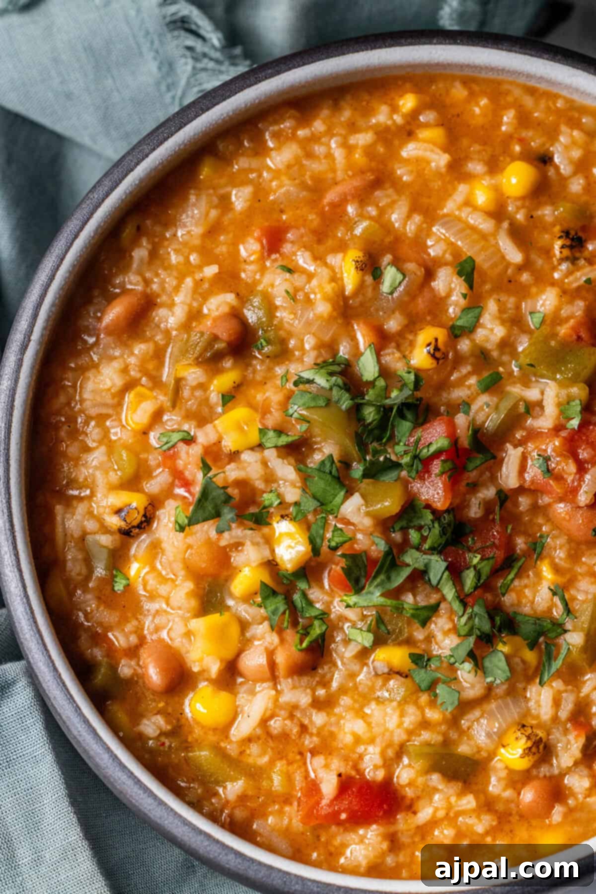 A bowl of vegan vegetable soup with rice, corn, beans, tomatoes, and green herbs, served on a light blue cloth.